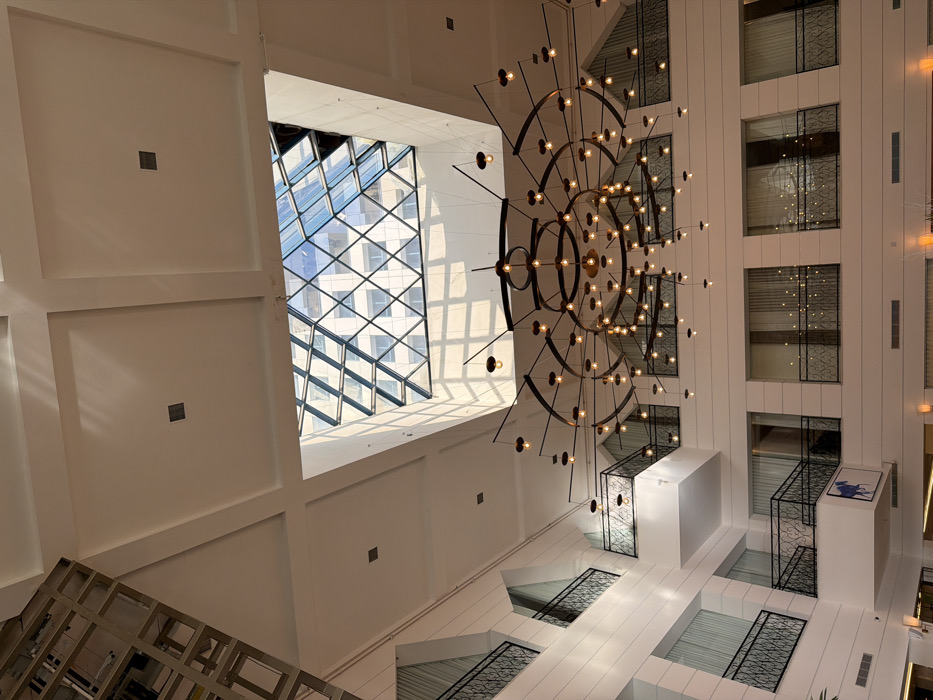 The image displays the multi-story atrium lobby of the Dan Tel Aviv Hotel, located in Tel Aviv, Israel, viewed from a low-angle perspective looking upwards. A large, elaborate chandelier, constructed from dark metal with multiple concentric rings and radiating arms, is centrally suspended from the high, white, recessed panel ceiling. Numerous small, exposed filament light bulbs are affixed to the chandelier. Directly above the chandelier, a large glass skylight, featuring a diamond-patterned grid, allows natural light to penetrate the space, casting distinct geometric shadows on the upper walls and ceiling. The skylight reflects the exterior facade of an adjacent building characterized by grid-patterned windows.
The atrium walls are white with horizontal paneling, and multiple levels of balconies or walkways are visible, each equipped with ornate black wrought iron railings featuring scrollwork designs. Behind these railings, windows or openings to rooms are apparent, some covered with sheer white curtains. On a wall of a lower balcony, a stylized blue abstract artwork, resembling a horse, is mounted. In the upper left portion of the frame, a partial view of a metallic, angular structural or decorative element is visible. No individuals are present in the scene. The lighting indicates daytime conditions.
