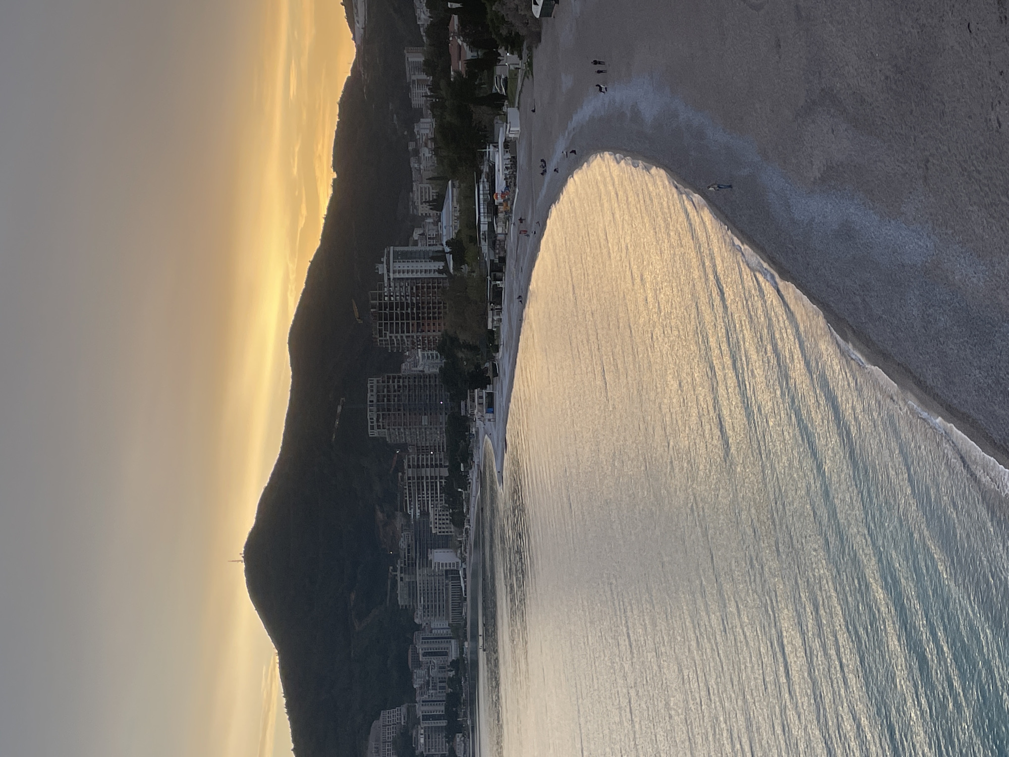 The image depicts a coastal scene in Budva, Montenegro, possibly taken during sunset or sunrise. A mountain rises to the left side of the frame, alongside a line of buildings including apartments, hotels, and other structures, extending along the coastline. A beach curves along the water's edge. The water surface has a reflective pattern, possibly from the sun's rays. Several small figures of people are visible on the beach and in the water. The sky is pale with a gradient of color from yellow to grey, indicating the presence of sunlight at a low angle. A tower is visible at the peak of the mountain.