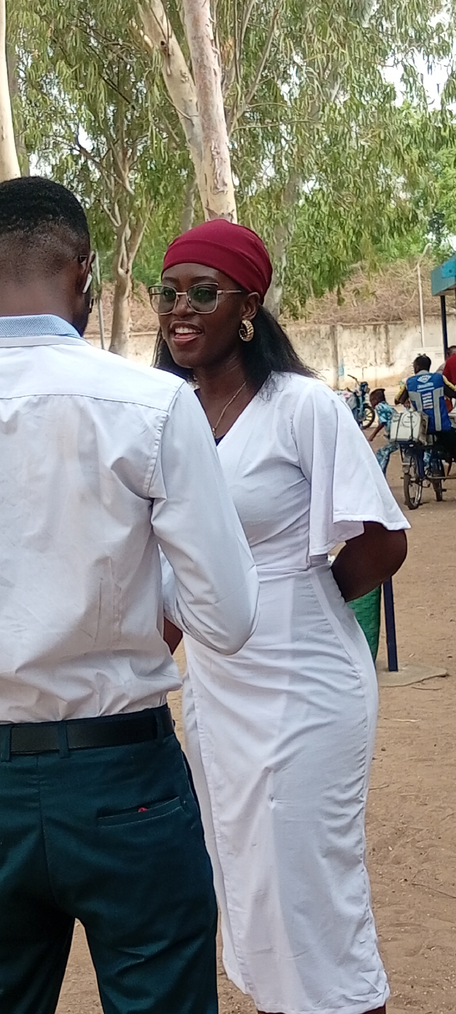The image shows two people in an outdoor setting in Fagge D2, Nigeria. On the left, a man is seen from the back, wearing a white shirt and dark green pants with a black belt. He has short black hair and appears to be wearing glasses or earphones. To the right, a woman is facing him, wearing a white dress, a burgundy head covering, and gold hoop earrings. She has dark hair and is wearing glasses. Both seem to be engaged in a conversation. In the background, there are trees, a building, and other people, including one person riding a bicycle. The ground appears to be a dusty or sandy surface.