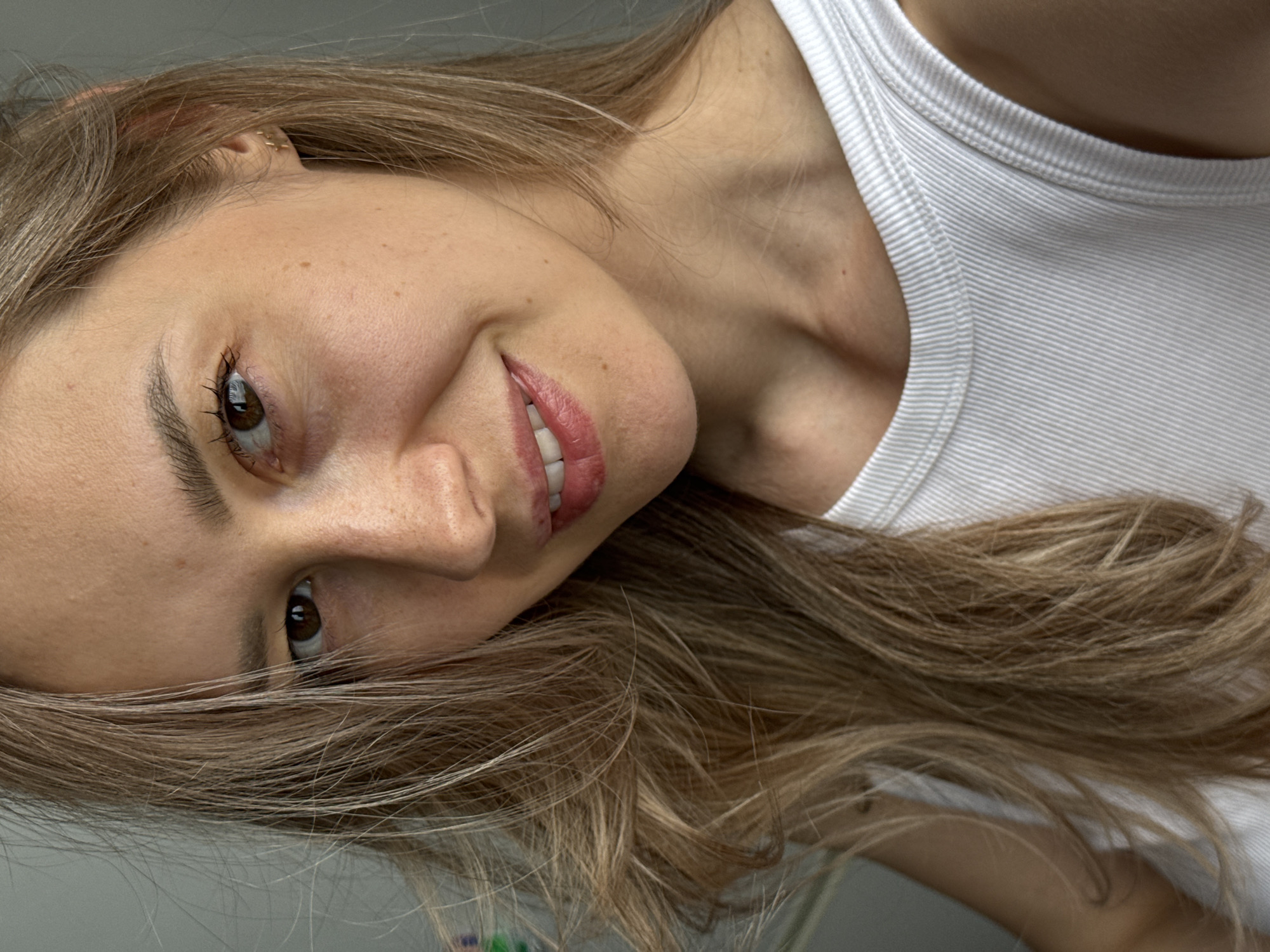 A woman with light brown hair and fair skin is smiling in an indoor setting. She is wearing a white ribbed tank top. A small gold hoop earring is visible in her left ear. The image does not provide any information related to the city of Chaikovsky, Russia.