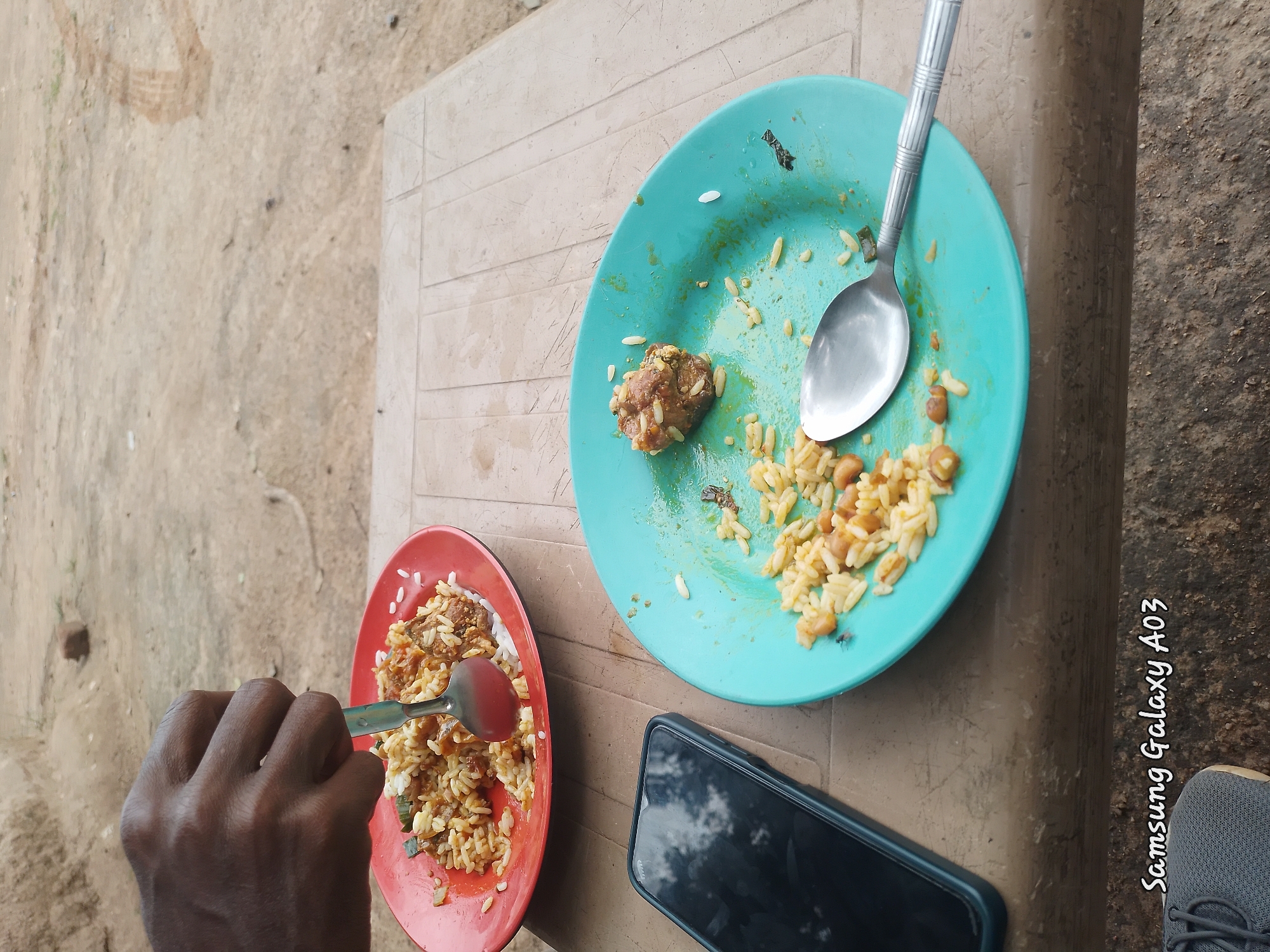 A close-up shot captures a hand using a spoon to serve food from a red plate. The plate is filled with rice and some meat. Next to it, a turquoise plate contains remnants of rice and a piece of meat, along with a spoon. A black smartphone rests on a textured table. The background shows a sandy, outdoor environment. The overall scene suggests a meal in progress or recently concluded in an unspecified location.