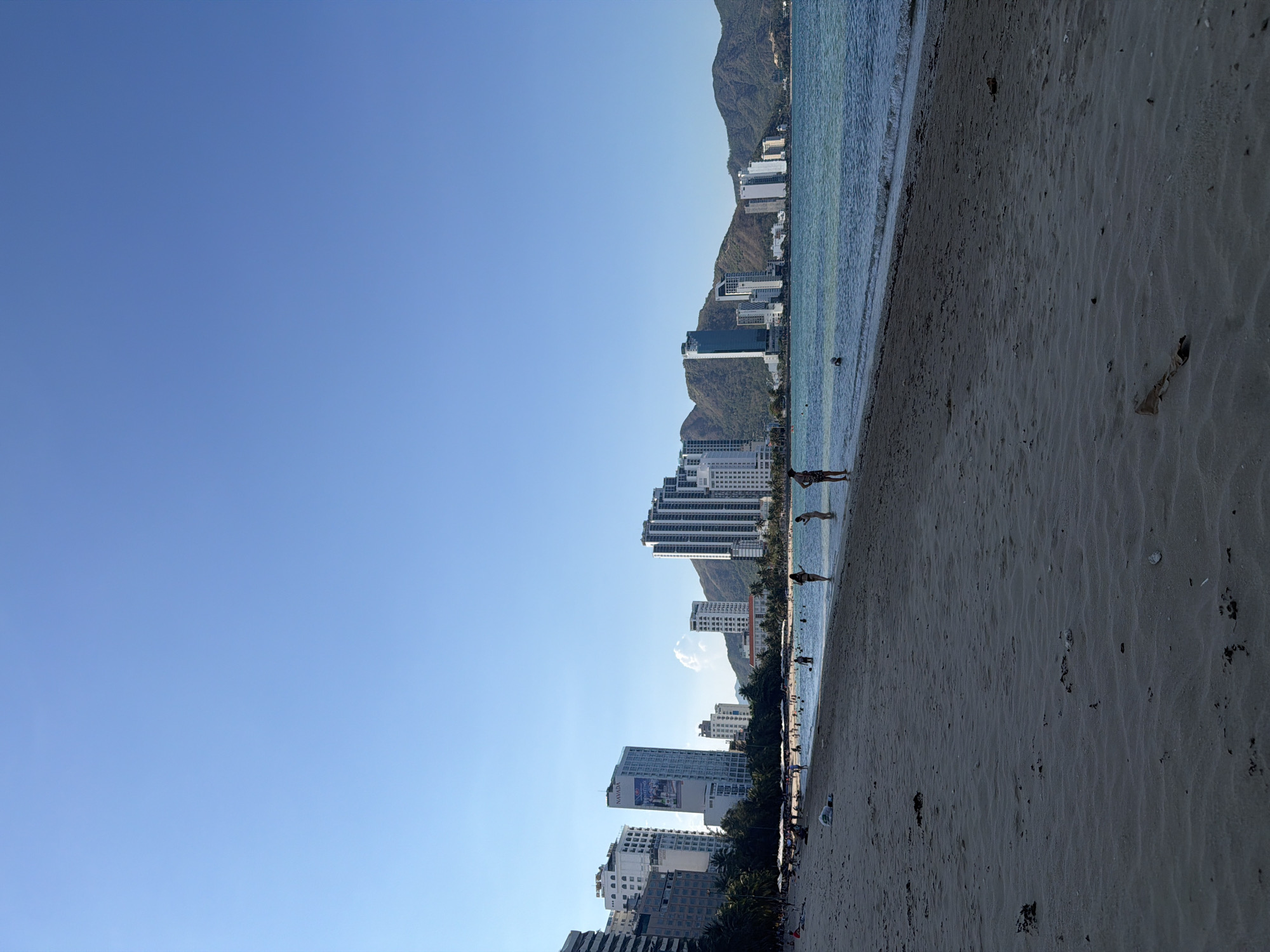 The image depicts a beach scene in Bac Nha Trang Ward, Vietnam. A clear blue sky fills the left half of the frame. The right half of the image showcases a sandy beach leading to the ocean. Several high-rise buildings are visible along the coastline, backed by mountains. People can be seen on the beach and in the water. The foreground of the beach shows footprints and scattered debris on the sand. The image appears to be taken during daylight hours with a clear sky.