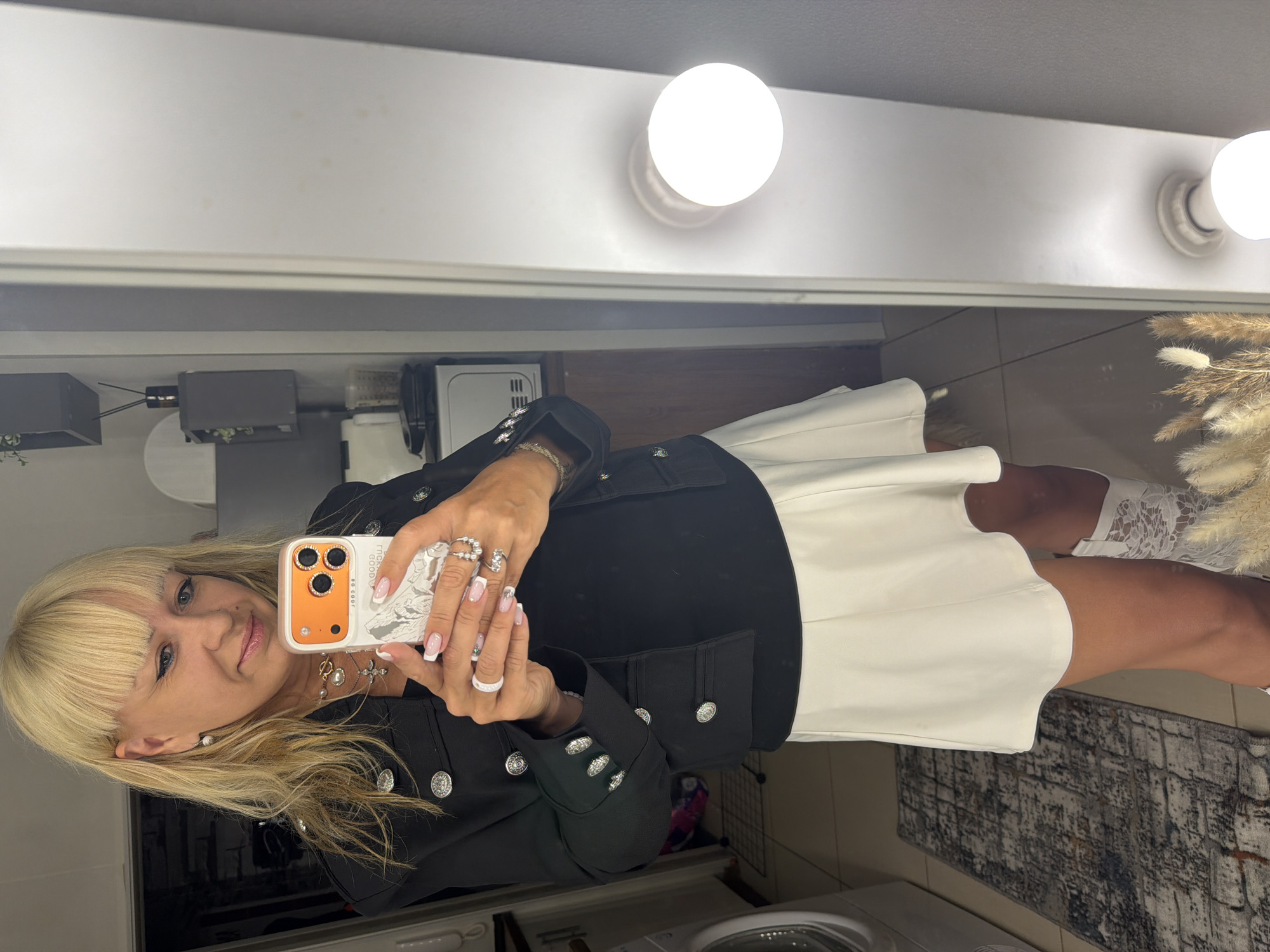A woman is taking a selfie in a room with a vanity mirror that has lights on the top. The woman has blonde hair with bangs, and is wearing a black jacket with silver buttons, a white skirt, and white lace socks. She is holding an orange phone with a white case in her hands. The phone case has the words "LOOK GOOD" written on it. The woman is also wearing multiple rings on her fingers and a necklace.
Behind her is a washer machine. Next to her is a light color rug. There is a plant next to the mirror.
The location is Russia, Chaikovsky.