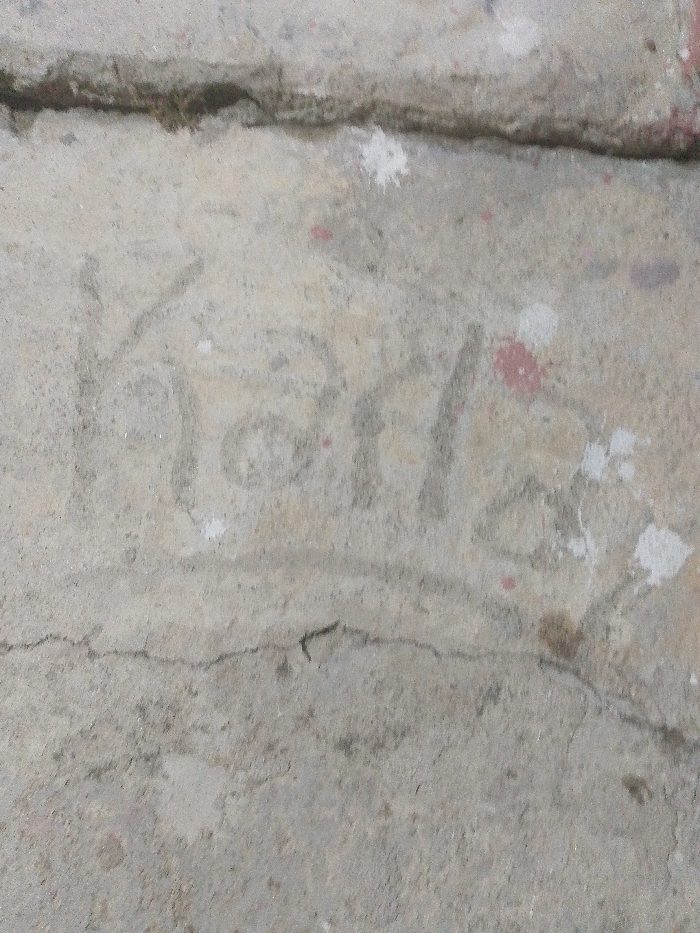 Faintly etched into a weathered, light gray concrete surface is the name "Karla." The lettering is thin and somewhat uneven, suggesting it was scratched or drawn with a tool. The concrete shows signs of age and wear, with cracks and small splatters of white and red paint scattered around the inscription. The surface itself is slightly rough and textured.