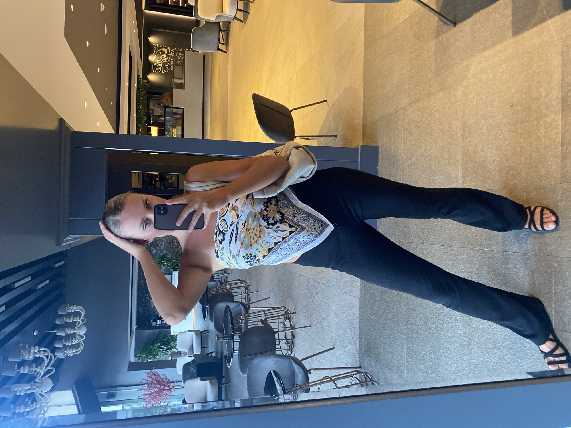 An indoor shot captures a blonde woman in her early 20s taking a selfie in a mirror. She is wearing a black strapless top, a patterned silk scarf tied around her chest, and black flared pants. She has her right hand in her hair and is holding her phone with her left hand. She is wearing black strappy sandals. She is in a lobby area that appears to be a hotel or cafe. The background shows seating areas with chairs and tables, and a counter area with food displays. The floor is tiled, and there is a large mirror reflecting the scene. The setting suggests a modern establishment, possibly in Aksu, Turkey, given the lighting and furnishings.