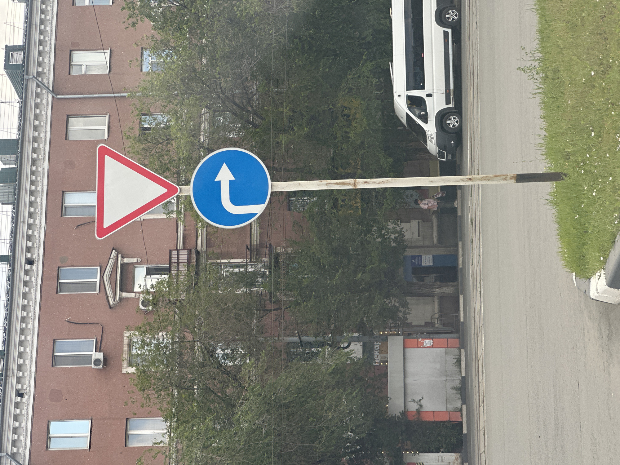 A vertical, medium shot captures a street scene in Orsk, Russia, featuring traffic signs, a building, trees, and a white van. In the foreground, a metal pole supports two circular traffic signs. The upper sign is a red-bordered inverted triangle, indicating "Give Way" (or "Yield"). Below it, a blue circular sign displays a white arrow pointing to the right, signifying a mandatory right turn.
Behind the signs, a multi-story brick building with numerous windows and air conditioning units is visible. Trees with green foliage partially obscure the lower sections of the building. To the right of the building, a white van is parked on the street. A lone pedestrian, a woman wearing a light-colored top and pants, stands near a bus stop, holding a bag. The street in front of the building is paved, with grass and wildflowers growing along the curb in the lower right corner of the frame. The overall lighting suggests an overcast day.