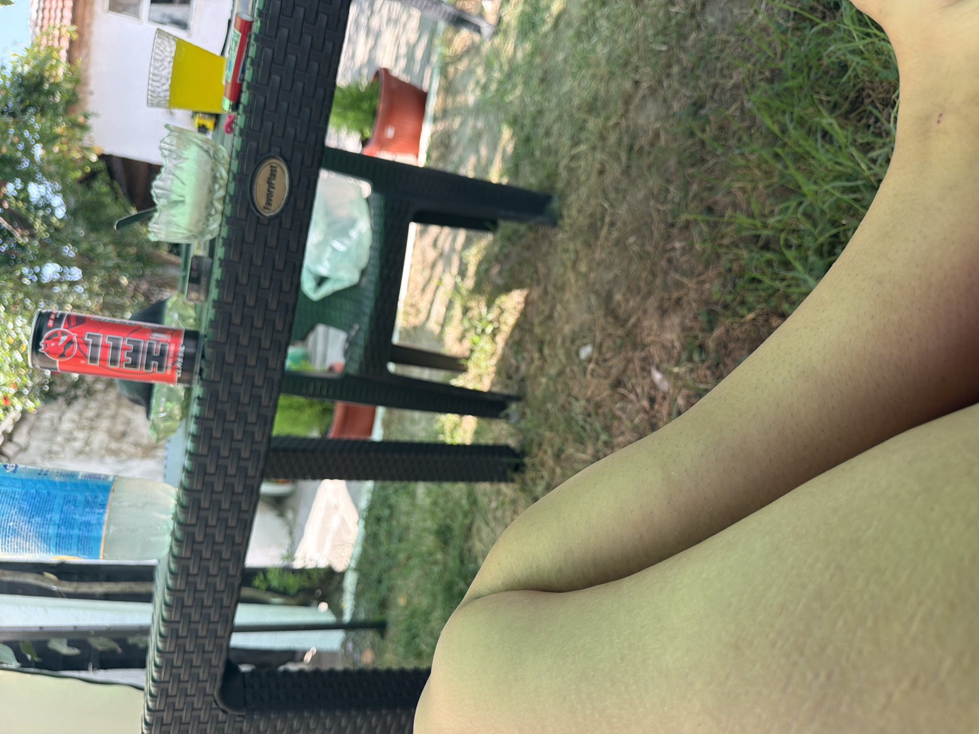 The image shows a person's legs resting on a grassy area in what appears to be an outdoor seating area. In the background, a woven plastic table holds several items, including a can of "HELL" energy drink, a glass bowl with what looks like sugar, a glass of yellow liquid, a cigarette pack, and a bottle of water. A plastic chair is partially visible behind the table, with a white plastic bag on it. The setting includes trees and a building in the distance, suggesting a residential area. The overall impression is a casual outdoor setting, possibly a garden or patio, in Razhevo Konare, Bulgaria, on a sunny day.