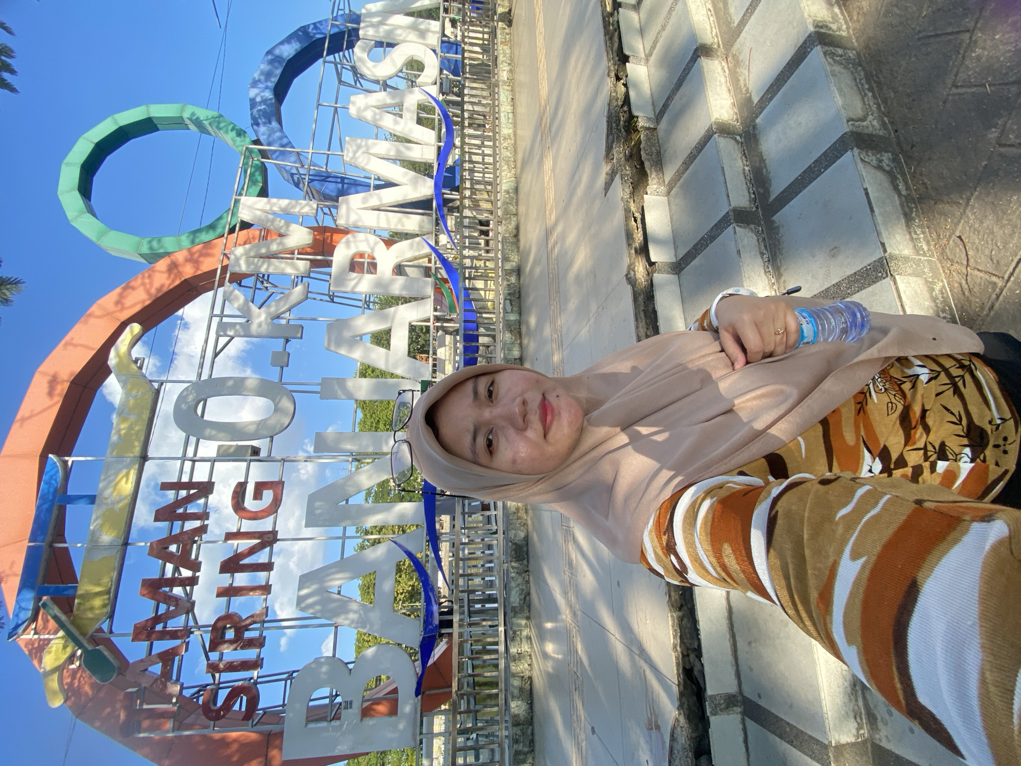 A young woman wearing a beige hijab and a patterned shirt takes a selfie in front of the "0 KM Banjarmasin" sign. The sign, large and colorful, features the words "AMAN SIRING 0 KM BANJARMASIN" in prominent white and red letters, set against a backdrop of blue sky and some clouds. The woman holds a bottle of water in her right hand. She is positioned in front of a paved area with steps leading up to the sign. The environment appears to be outdoors, possibly a public park or landmark, given the signage and open space. The overall scene suggests a tourist or visitor at a notable location in Banjarmasin, Indonesia.