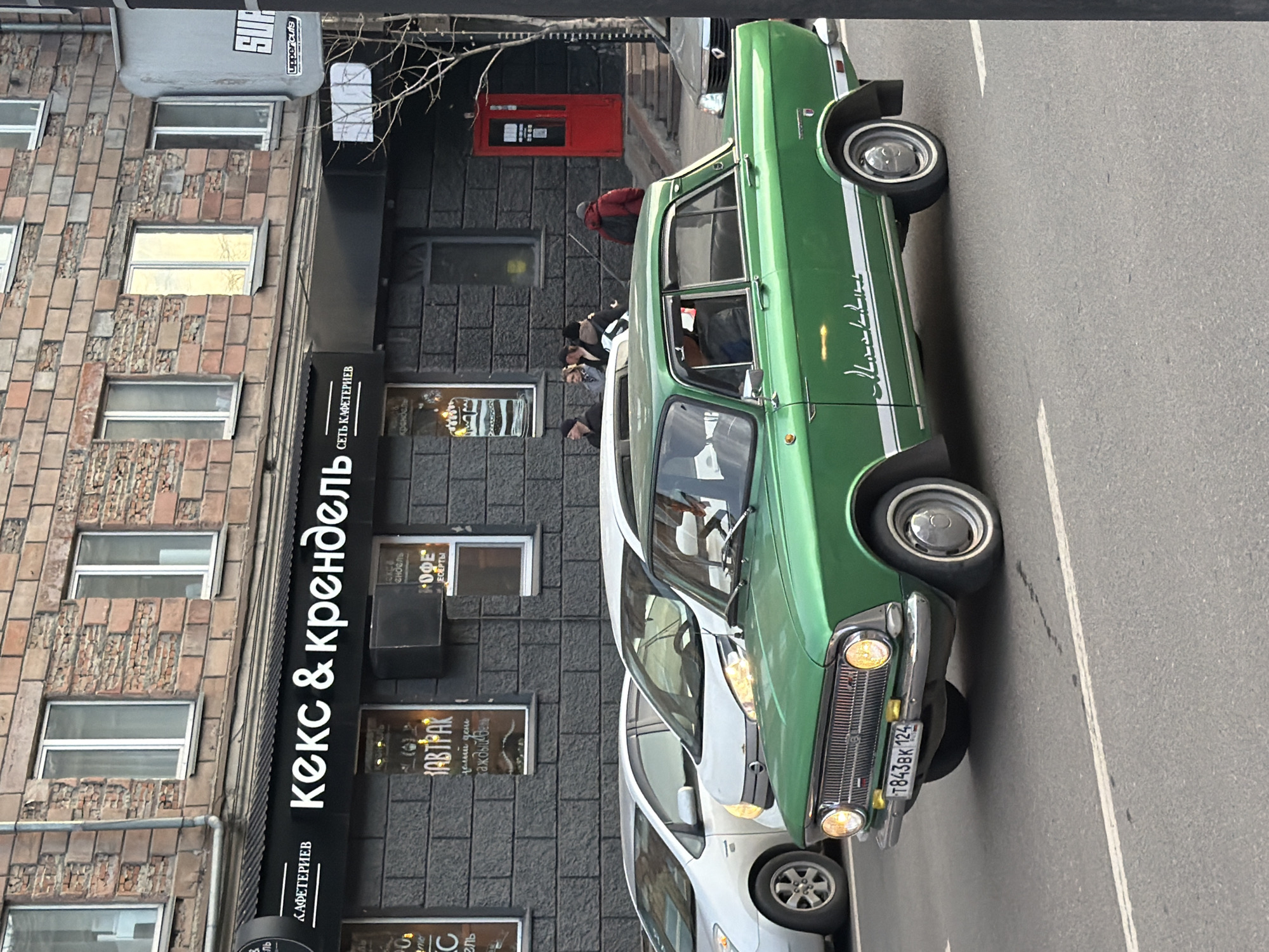 The image captures a rotated street scene, appearing as if viewed from above, with a building on the left and a road on the right. The setting is an urban environment, likely during an overcast day given the diffused lighting and lack of strong shadows.
The primary subject is a vibrant green classic car, possibly a Soviet-era model like a GAZ-24 Volga, parked along the side of the road. It features white racing stripes running horizontally along its body and has a chrome grille, round headlights, and a license plate that reads "Т843ВК 124". The number "124" signifies the Krasnoyarsk Krai region in Russia. Behind it, a modern white sedan is partially visible.
On the left, a multi-story building displays a facade of varying textures, including brickwork and rough stone sections, with numerous windows. Adjacent to this, a darker commercial building houses several establishments. Visible signage includes a prominent black awning with white Cyrillic text that reads "КЕКС & КРЕНДЕЛЬ" (Keks & Krendel, meaning "Cake & Pretzel" or "Muffin & Bagel"), and below it, "СЕТЬ КАФЕТЕРИЕВ" (Set' Kafeteriev, meaning "Chain of Cafeterias"). Other smaller signs in windows advertise "КОФЕ" (Kofe - Coffee), "ДЕСЕРТЫ" (Desserty - Desserts), "ЗАВТРАК" (Zavtrak - Breakfast), and "КАЖДЫЙ ДЕНЬ" (Kazhdyy Den' - Every Day). A red automated teller machine (ATM) or similar device is mounted on the dark building.
Several people are visible near the commercial storefronts; one person in a red jacket is seen near the ATM, and others are standing or walking further down. At the top left, a gray sign with "SUP" and "uppercuts" is discernible, suggesting another business. The overall scene depicts a typical street in a Russian city with commercial activity and parked vehicles.