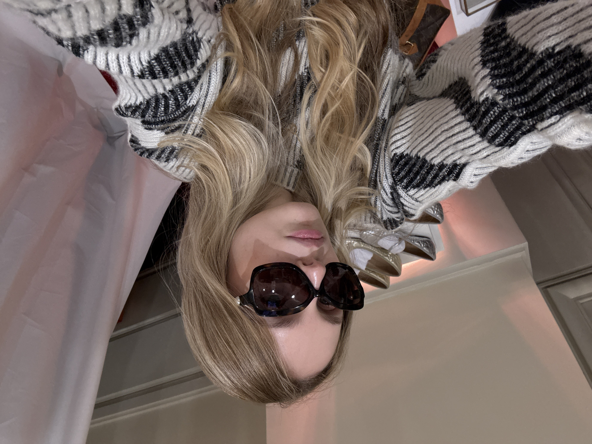 A woman with long blonde hair is lying on her back and looking directly at the camera. She is wearing large, dark sunglasses and a black and white patterned sweater. The background includes shoes on a shelf, a cabinet, and part of a white curtain. The location cannot be determined based on the image.