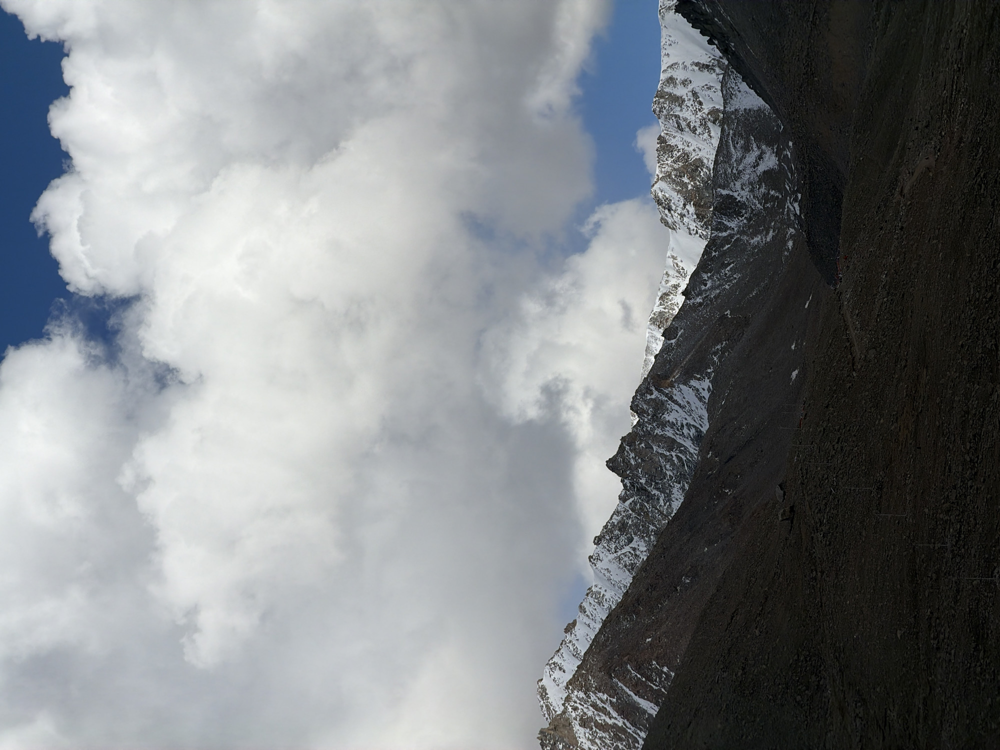 The image depicts a mountain range with snow-covered peaks on the right side of the frame. The other half is obscured by puffy white clouds. There are patches of blue sky visible. The mountain range is mostly dark with some patches of snow. No people, subjects, objects, actions, or interactions are observed. The location is not specified, it is a mountain range that contains snow and clouds.