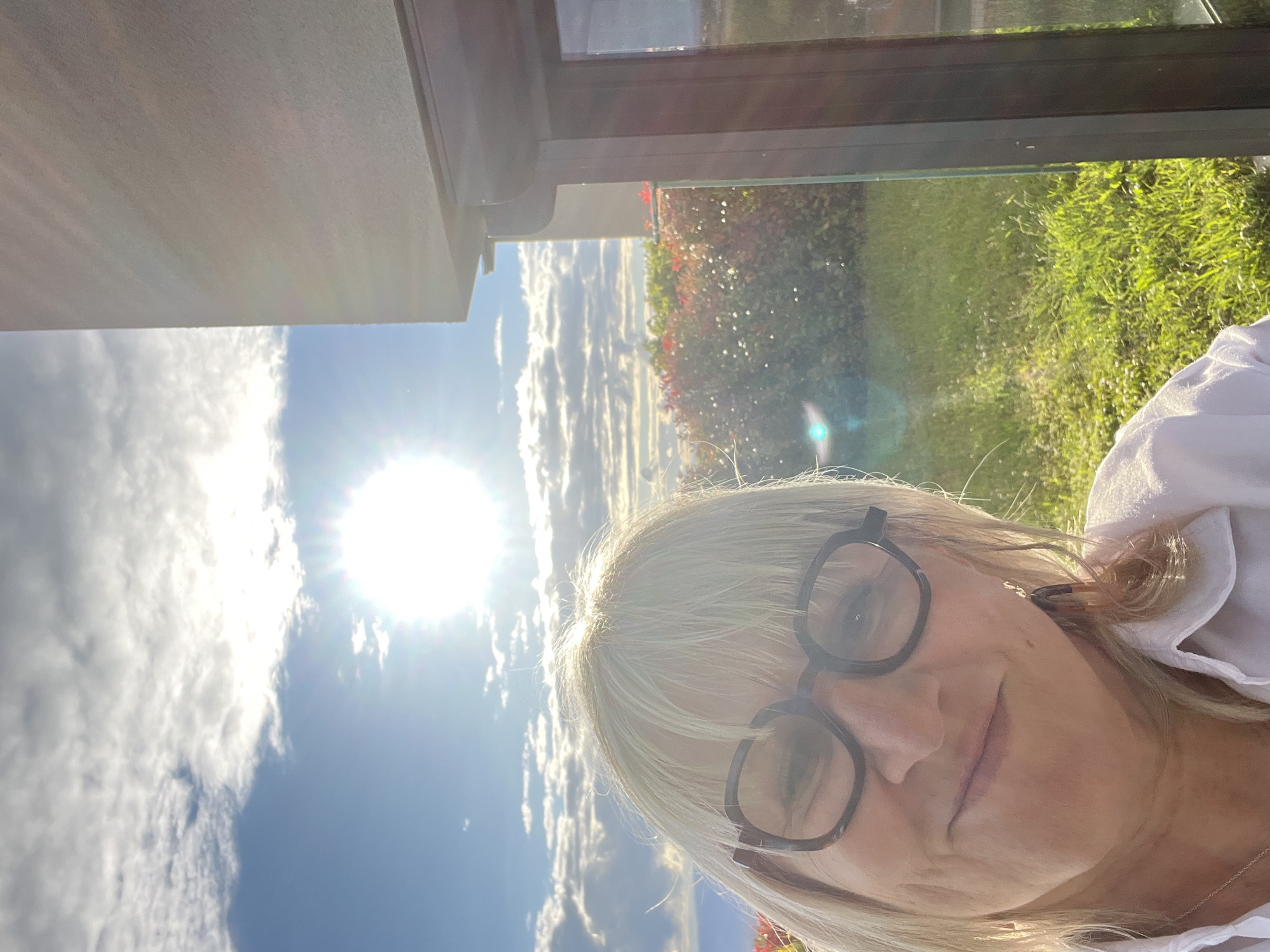 A close-up, slightly angled shot shows a blonde woman wearing dark-rimmed glasses. Her face is partially illuminated by sunlight. In the background, a bright sky with clouds is visible, along with greenery suggesting a garden or lawn. The top of a building or structure is also partially visible. The location is Chambœuf, France.