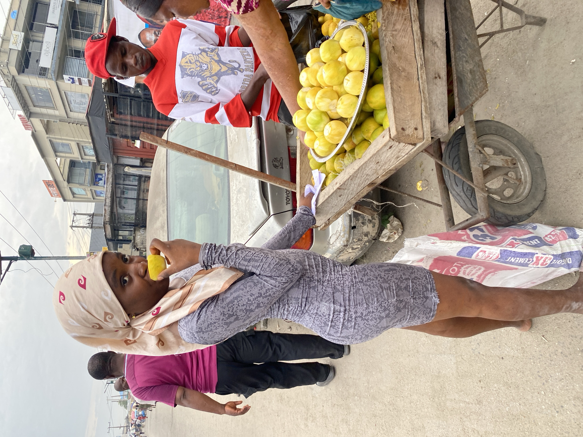 The image shows a woman eating a yellow fruit, likely a lemon or orange, while standing near a fruit vendor's cart in Lagos, Nigeria. The vendor, a man wearing a red and white hockey jersey, stands behind the cart which is filled with the yellow fruit. A white sedan is parked behind the cart. A man in a purple shirt walks past. The background shows a city street with buildings and overcast skies.