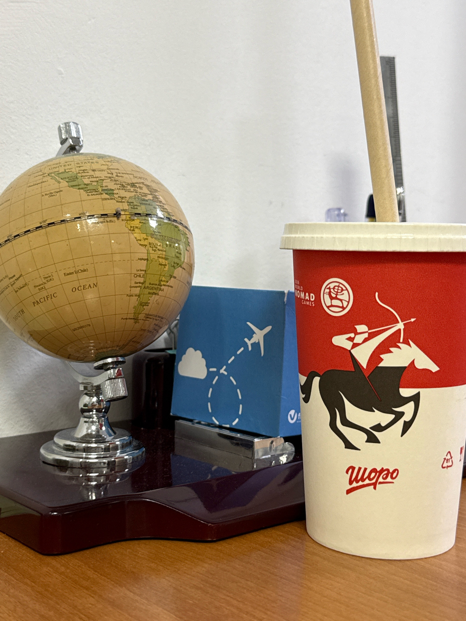 A desk in Bishkek, Kyrgyzstan features a vintage globe with visible country names such as "PACIFIC OCEAN" and "SOUTH AMERICA." To the right of the globe is a red and white paper cup with a depiction of a horse and rider, and a brand name "Wopo." A light-colored paper straw is inserted into the cup. Behind the cup, a blue box with a white airplane graphic is partially visible. The objects are situated on a dark, glossy surface, which rests on a wooden desk. The background is a plain, white wall.