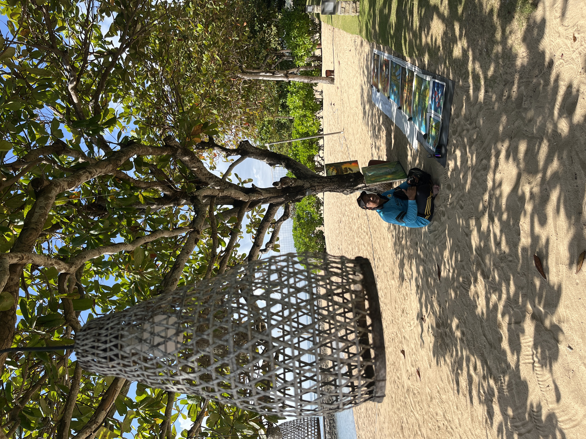 In Kuta Selatan, Indonesia, a person sits on a sandy beach under the shade of a tree, displaying several paintings laid out on a mat. The individual is wearing a blue long-sleeved shirt with yellow accents and dark pants, and appears to be a male. He is holding what looks like a camera or some kind of electronic device. In the foreground, a woven lantern hangs from a tree branch. The background shows a body of water, lush green foliage, and a volleyball net. The scene suggests an outdoor art display or market on a sunny day.