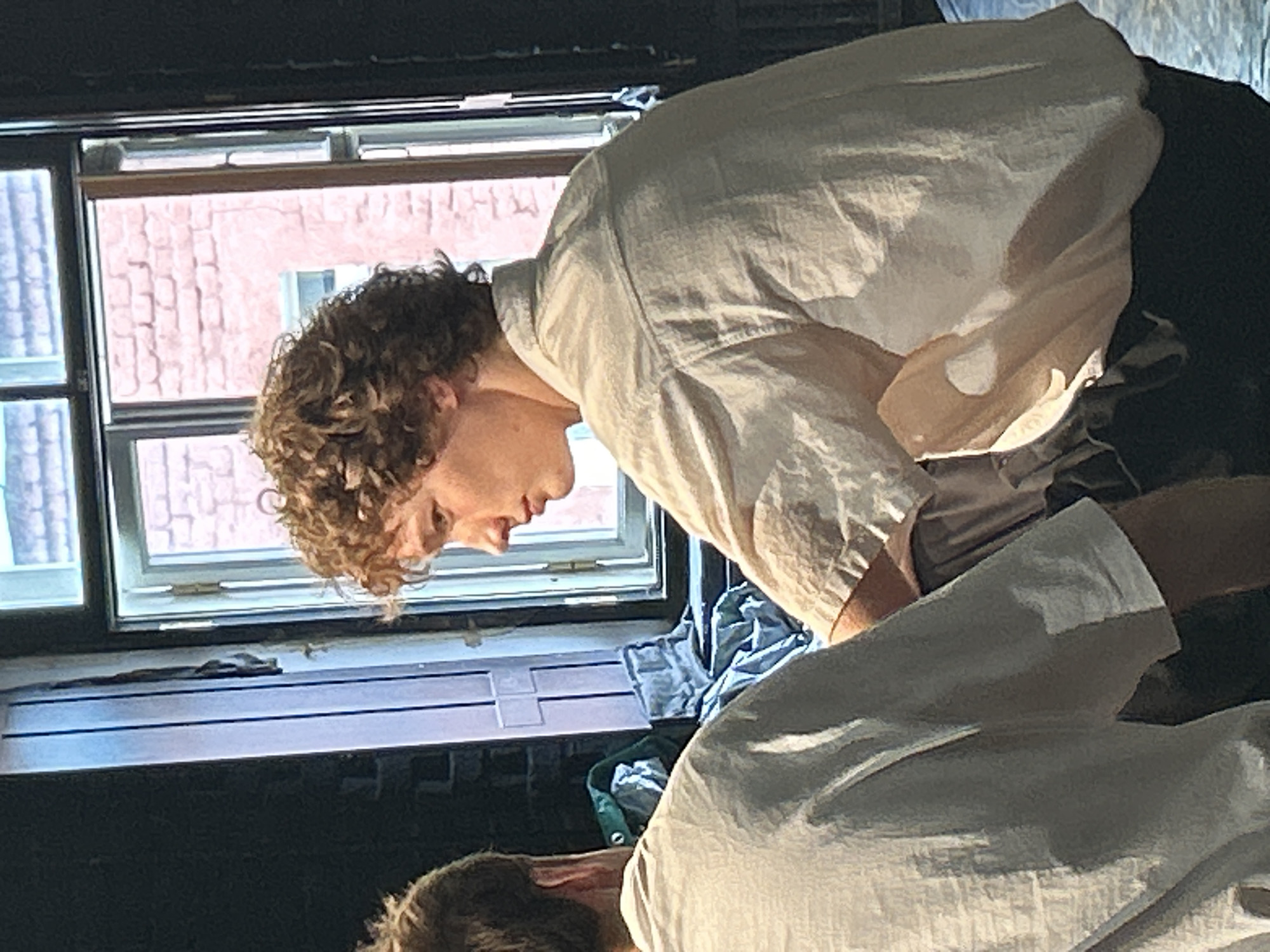 A young man with curly blonde hair, wearing a light-colored, loose-fitting shirt, is the central subject of the image. He is positioned in profile, facing to the left, with his gaze directed downwards. His facial features are distinct, with a prominent nose and slightly parted lips. He appears to be in a seated or crouched position.
Behind him, to the left, is another person, seen from the back and shoulder, also wearing a light-colored shirt. This individual's head is turned away from the camera, and their hair is short and dark.
The environment suggests an indoor setting, possibly a room with a large window. Through the window, a brick building is visible, indicating an urban location. The window frame is visible, with multiple panes, and appears to be a casement window, slightly ajar. The interior includes dark walls and what appear to be stacks of fabric or clothing in muted colors. The lighting is soft, with natural light streaming in from the window, casting shadows and highlighting the textures of the clothing. The overall atmosphere is somewhat informal and relaxed, possibly a rehearsal space or a casual gathering. The specific city and location within the city are unknown.