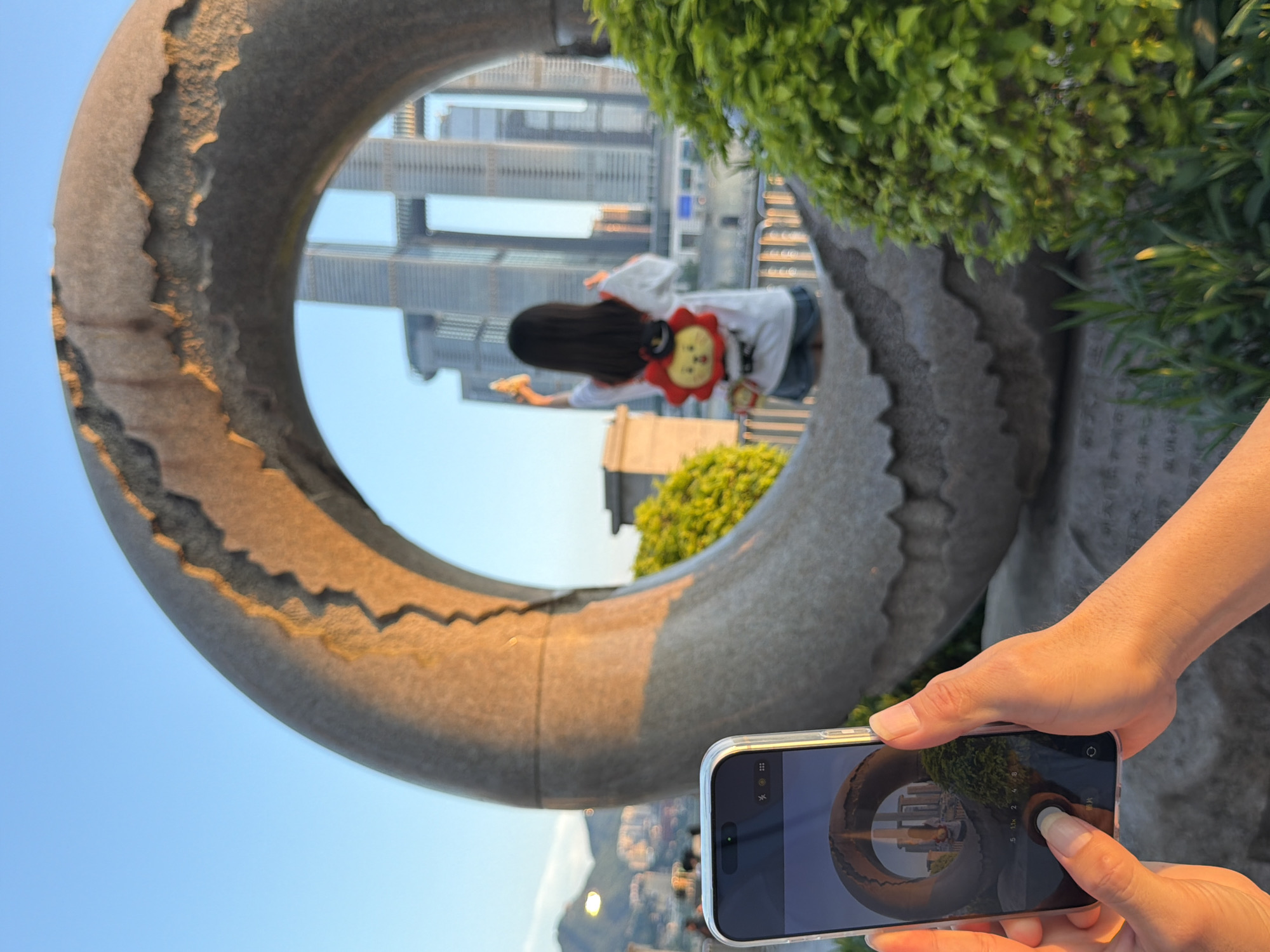A person holds a smartphone in the foreground, capturing a photo of a circular stone sculpture. The sculpture frames a view of a young person, seen from behind, wearing a white shirt, denim shorts, and a backpack shaped like a lion's face. This person is holding up an ice cream cone and appears to be standing near a river or waterfront. In the background, towering modern skyscrapers dominate the skyline, indicative of an urban environment. The scene is bathed in the warm light of what appears to be late afternoon or early evening, casting golden hues on the stone and foliage. The presence of skyscrapers and the overall urban setting suggests this photo was taken in a major Chinese city. Given the context provided, the location is identified as Nan'an District, China.