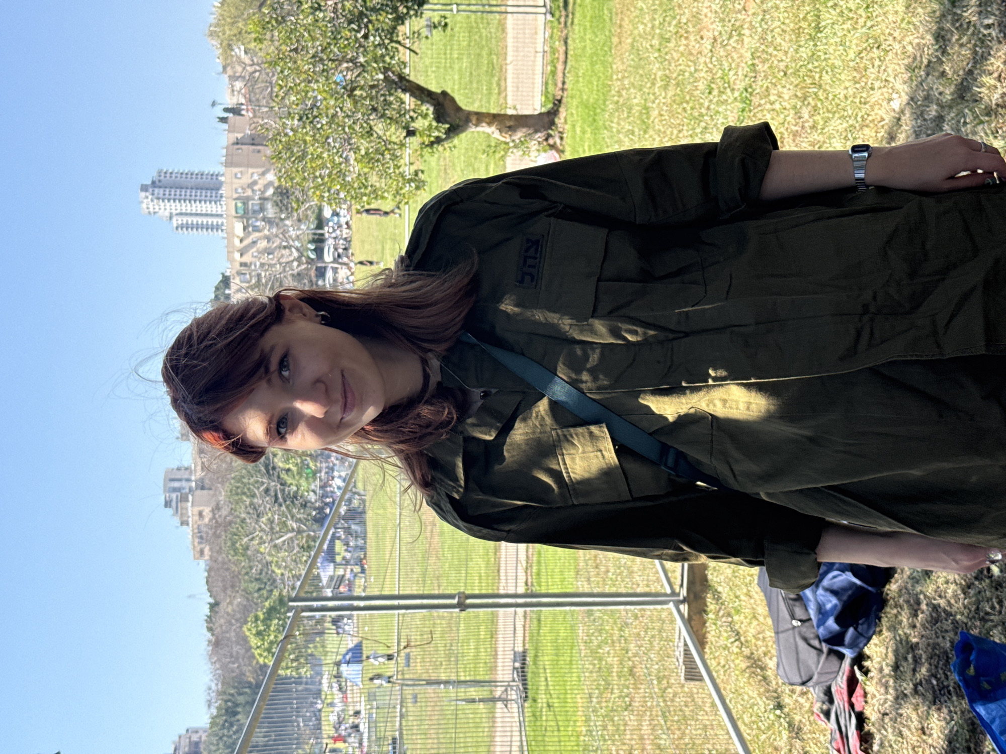 A young woman in an olive-green military uniform is standing in a park on a sunny day. Buildings can be seen in the background, suggesting a city location. The woman has fair skin, auburn hair, and is wearing a watch. She appears to be posing for the photo. The uniform has a patch with Hebrew lettering, likely identifying her as a member of the Israel Defense Forces (IDF). A blue strap is visible across her chest. The park setting includes green grass and trees, as well as a metal fence. In the background, a crowd of people and vehicles are observed. Considering the architectural style of the buildings and the language on the uniform, the location is likely in Tel Aviv, Israel.