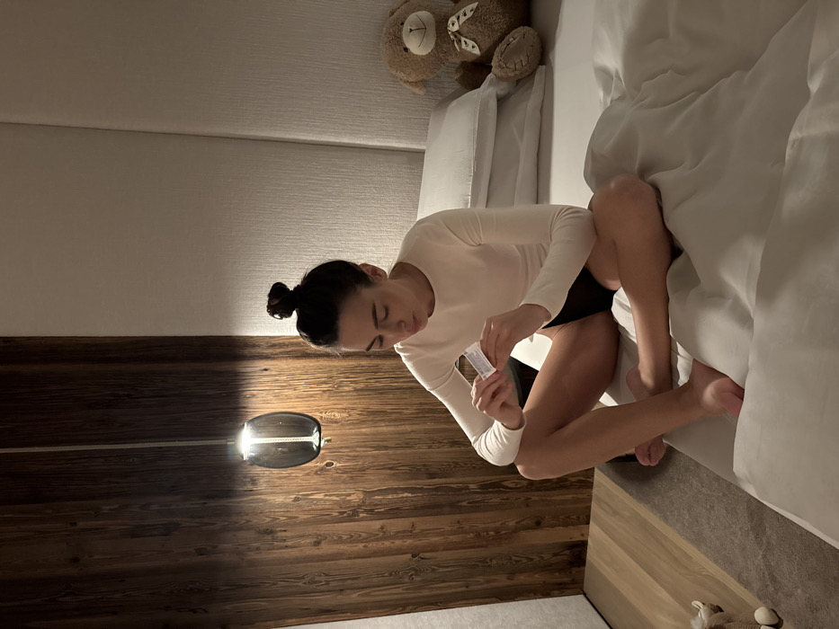 The scene is an interior room, consistent with a modern bedroom or hotel room, located in Kastelruth - Castelrotto, Italy. A female individual with dark hair styled in a top knot is seated cross-legged on a bed, facing downwards and slightly to the right of the frame's center. She wears a long-sleeved, off-white top and dark briefs. Her hands hold a small, white, rectangular object, which she is intently examining. The bed is covered with white bedding and features multiple white pillows against a light-colored, possibly upholstered, headboard on the right. A brown teddy bear with a light-colored ribbon is positioned on the bed to the right of the individual. To the left, a wall section is paneled with dark, natural wood. A modern pendant light fixture with a translucent dark grey glass shade hangs from the ceiling in front of the wood paneling, emitting a soft light. The floor is covered by a grey carpet or rug, with a small, light-colored stuffed animal partially visible in the lower left corner. The room is dimly lit, with illumination primarily from the pendant fixture and ambient light.