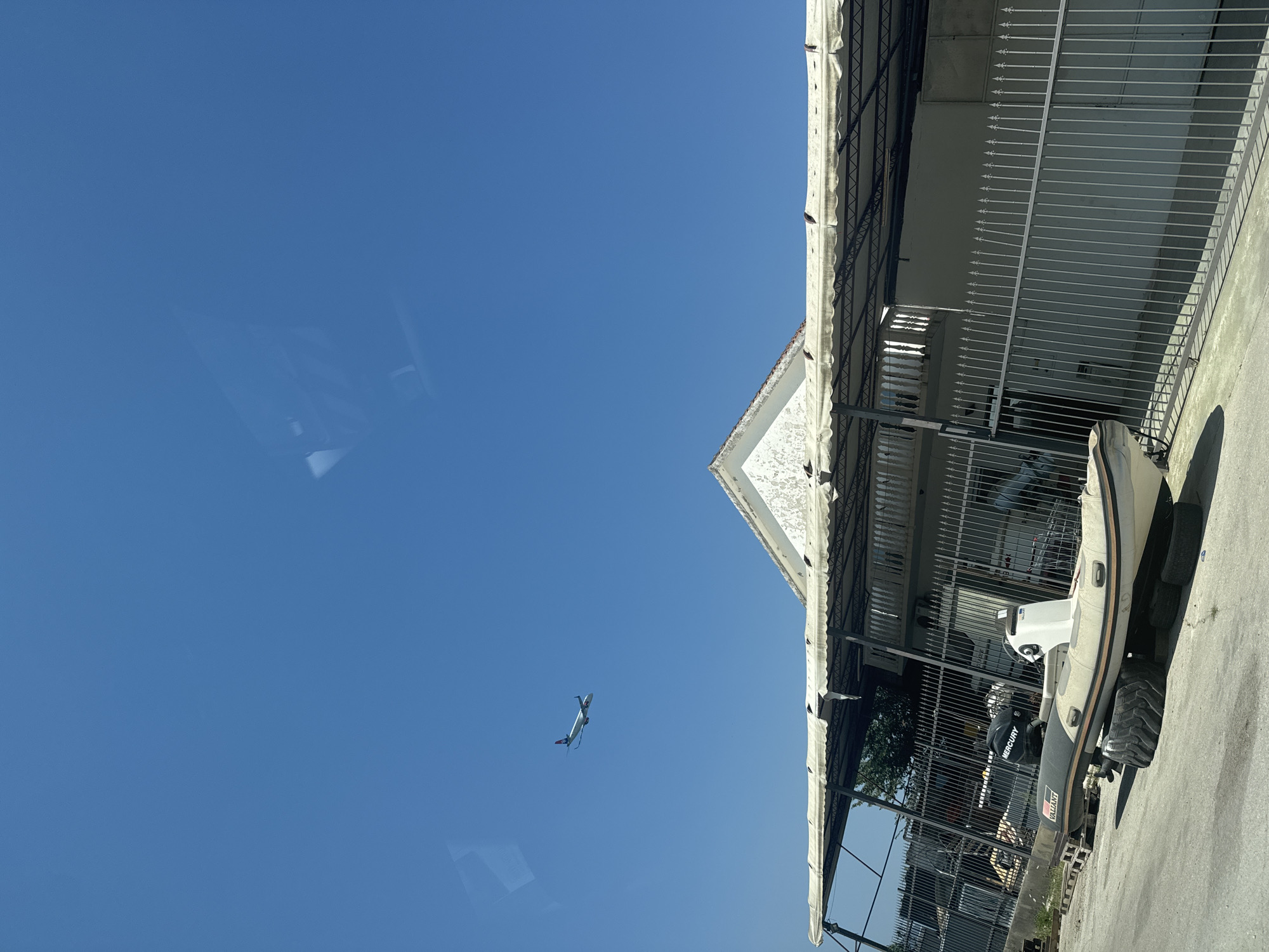An aerial view captures a clear blue sky, with a commercial airplane flying in the upper left quadrant. Below the sky is a white building with a peaked roof, partially obscured by an awning. A metal fence runs horizontally across the midground, separating the foreground from the building. In the foreground, a rigid inflatable boat rests on tires. The scene appears to be an outdoor storage or workshop area. The location is identified as Mrčevac, Montenegro.