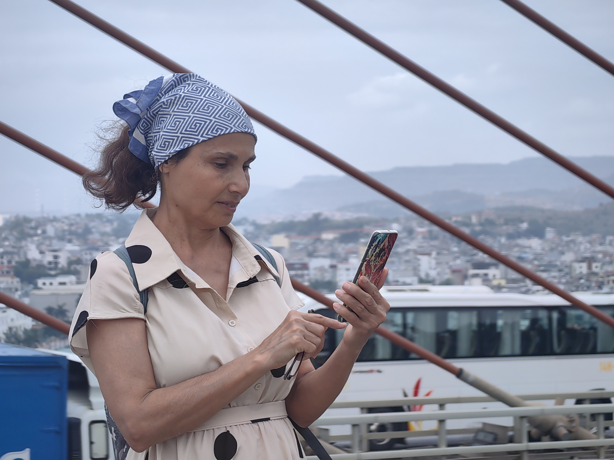 A woman is captured in a medium shot, standing outdoors and intently engaged with her smartphone. She is positioned slightly to the left of the frame, facing towards the right with her head tilted downwards as she looks at her device. She wears a blue and white patterned headscarf, tied at the back, covering her dark hair which is styled in a small ponytail. Her attire consists of a collared, short-sleeved, beige or cream-colored dress adorned with large black polka dots. A narrow belt cinches her waist, and a backpack strap is visible over her left shoulder.
Her right index finger is extended, poised to interact with the colorful screen of her smartphone, which she holds in both hands. The phone is encased in a vibrant, multi-colored case featuring intricate, possibly floral or abstract patterns. In her left hand, she also holds a pair of dark-rimmed glasses. Her expression is focused and neutral, indicating concentration on her phone activity.
The background presents an urban landscape under an overcast, light gray sky. Several prominent reddish-brown diagonal beams traverse the frame, suggesting a bridge or architectural structure. Beyond these beams, a cityscape with various buildings is visible, and in the far distance, hazy mountains rise against the horizon. A white bus with dark windows is seen in the mid-ground, along with a white railing. On the left side of the frame, part of a blue container or vehicle is present. The scene depicts a daytime setting in what is known to be Bai Chay Ward, Vietnam. No clearly legible text is visible on any object.
