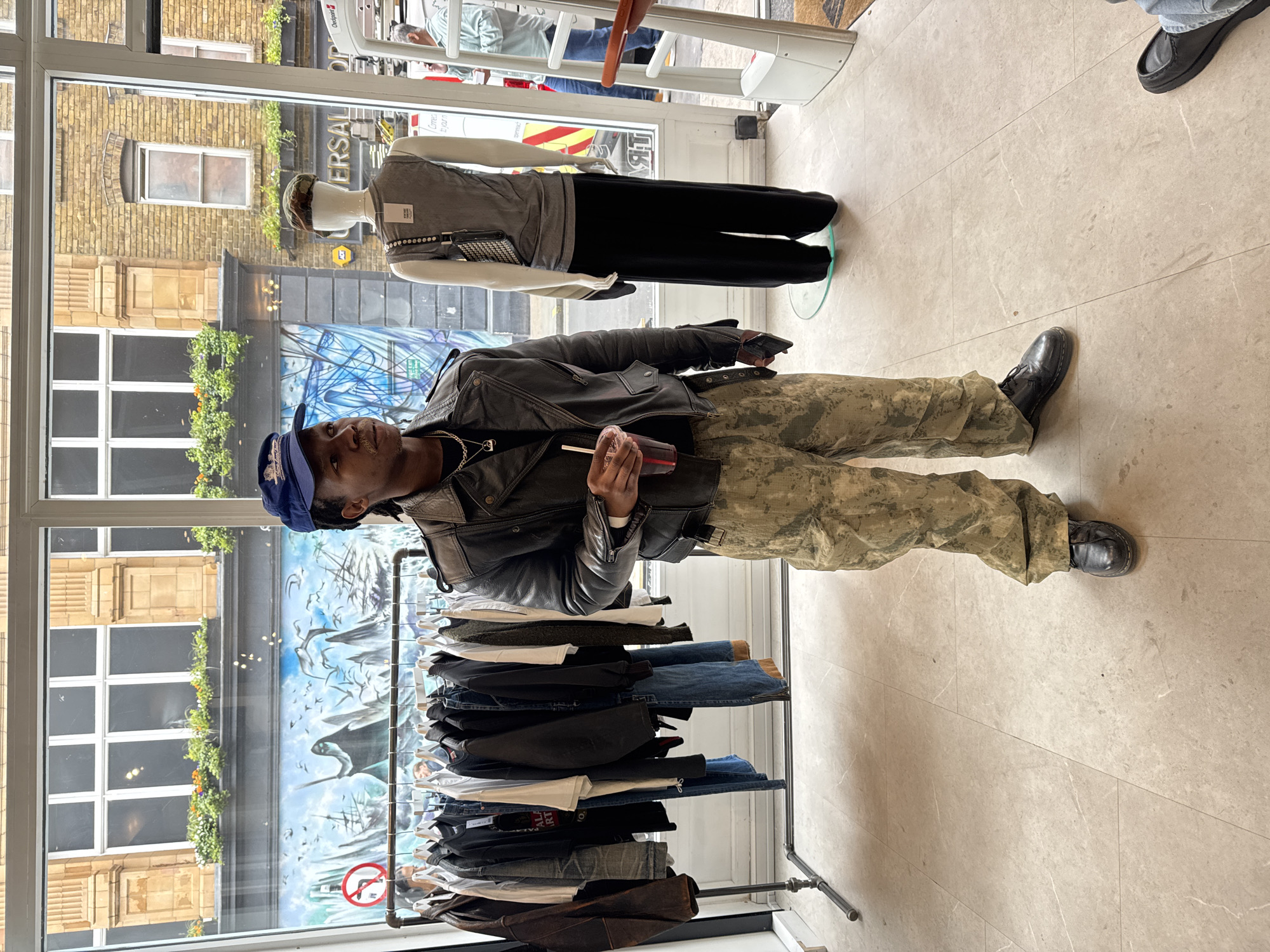 The image shows a full-length view of a young Black man standing inside a retail store, looking slightly towards the camera. He is wearing a blue baseball cap, a black leather jacket, camouflage cargo pants, and black boots, with a silver chain visible around his neck. He holds a clear plastic cup with a red straw in his right hand.
To his left, a metal clothing rack displays various garments, including jeans, t-shirts, and jackets. On the right, a headless mannequin stands on a circular clear base, dressed in a grey sleeveless top, black trousers, and a dark patterned hat, with a small black shoulder bag draped over its arm. The store floor is made of light-colored, large square tiles.
The setting is indoors, but a large window behind the man reflects an urban street scene. Reflections show a multi-story brick building with windows, some featuring window boxes with greenery and flowers. A large mural or artwork on an opposing building depicts birds flying over stylized mountains or ships, featuring a prominent red circular "no smoking" sign. Other reflected signs include "UNIVERSAL" and a yellow "AA" logo. A "OnePoint" sign is also visible. Partially visible through the window and inside the store are other individuals; a person's feet in blue jeans and black shoes are seen in the upper right, and another person is partially obscured by a white railing further back. The scene is illuminated by natural daylight.