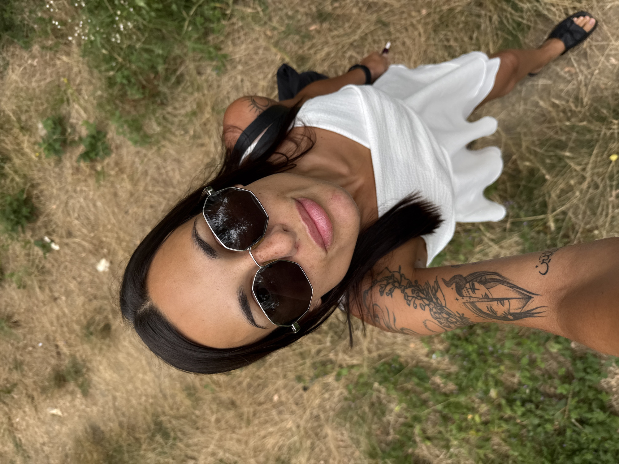 The image shows a woman taking a selfie outdoors. She is wearing a white dress and sunglasses. Her left arm is visible, and it has tattoos. The background appears to be a grassy or dry field with some vegetation. The setting does not provide any specific clues about the location within the city of Pleven, Bulgaria.