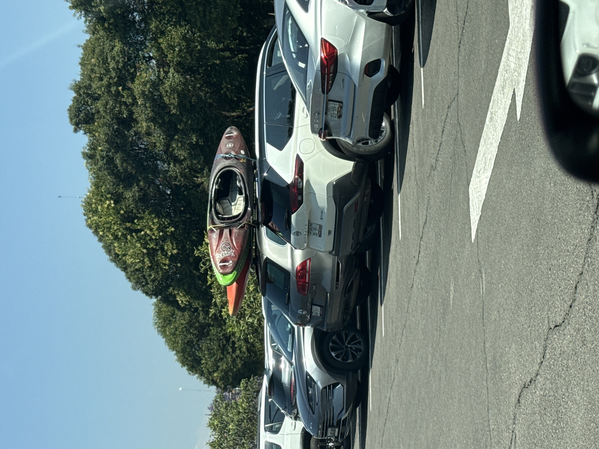 The image captures an outdoor parking lot scene on a clear, sunny day. Several vehicles are parked in a row, with a line of dense green trees in the background under a bright blue sky.
In the foreground, a white SUV is prominently featured, equipped with a roof rack that securely carries a reddish-brown and green kayak. The kayak has the word "Outbound" clearly visible on its side. Behind this vehicle, another light grey SUV is parked, displaying a Mexican license plate with the alphanumeric sequence "L-19-SFY-8". A second white SUV, partially obscured by the kayak's front, also has a visible Mexican license plate showing "MEXICO" at the top and the numbers "478-43" at the bottom.
The parking lot surface is asphalt, marked with white painted lines delineating parking spaces and a large white directional arrow. Minor cracks are visible in the asphalt. A small, faint white object can be seen high in the distance against the blue sky. The scene depicts a moment in an outdoor parking area, likely during the day due to the strong sunlight and clear visibility. The kayak suggests an ongoing or planned recreational activity. The overall setting aligns with an urban or suburban environment in San Andrés Cholula, Mexico.