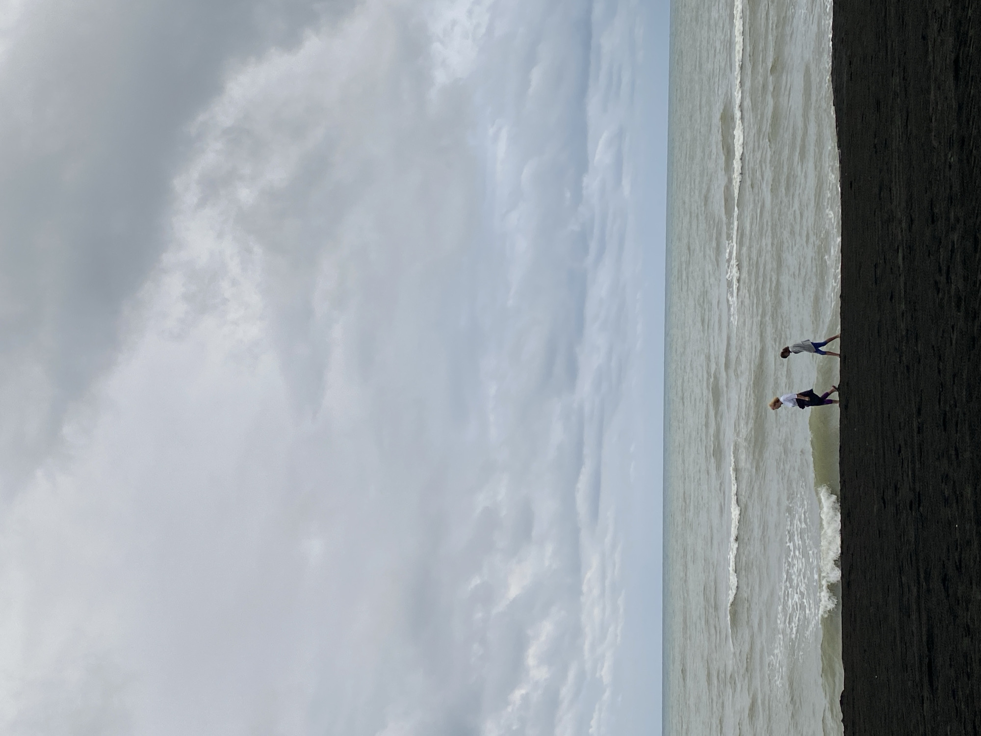 Two individuals are walking along a dark, sandy beach in Ureki, Georgia. The person on the left is wearing a white shirt and dark pants, and the person on the right is wearing a gray hooded jacket and blue pants. They are walking parallel to the water, which has moderate waves. The sky is overcast with dense, gray clouds. The overall scene depicts a somewhat somber, natural environment.