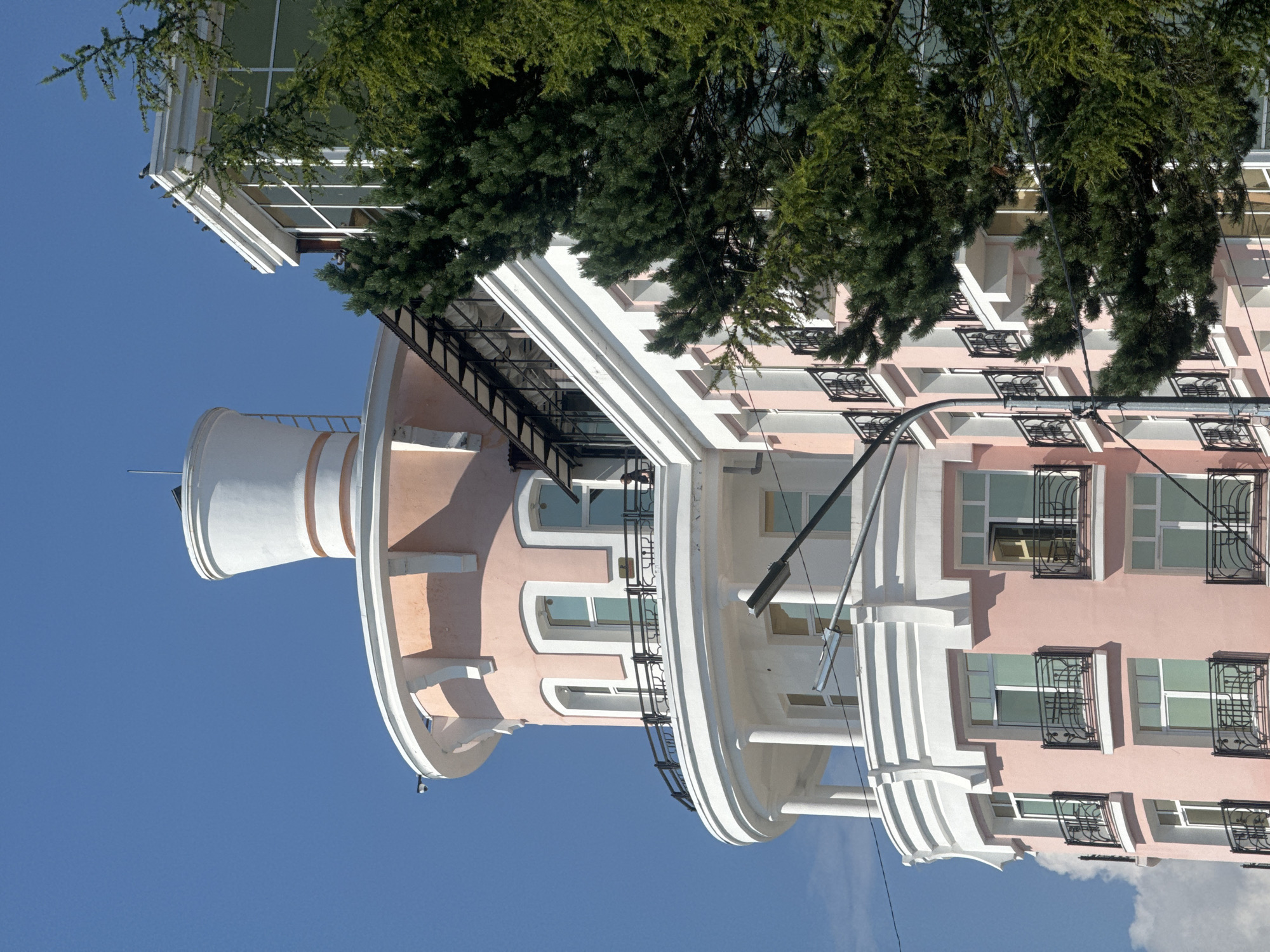 The image captures a close-up view of a pink and white building with a rounded facade and a distinctive white cylindrical top structure against a clear blue sky. The building features multiple floors with white-framed windows, each equipped with black wrought-iron balconies. A prominent streetlamp extends into the frame from the lower right. To the right of the building, a large green coniferous tree partially obscures the view. The scene appears to be an urban or semi-urban setting, given the presence of the building and streetlamp. The architectural style suggests a modern or renovated structure with some decorative elements. The overall impression is one of a bright, sunny day. The location is identified as Listvyanka, Russia, a settlement on the southwestern shore of Lake Baikal, known for its scenic views and wooden architecture, though this building exhibits a more contemporary style.