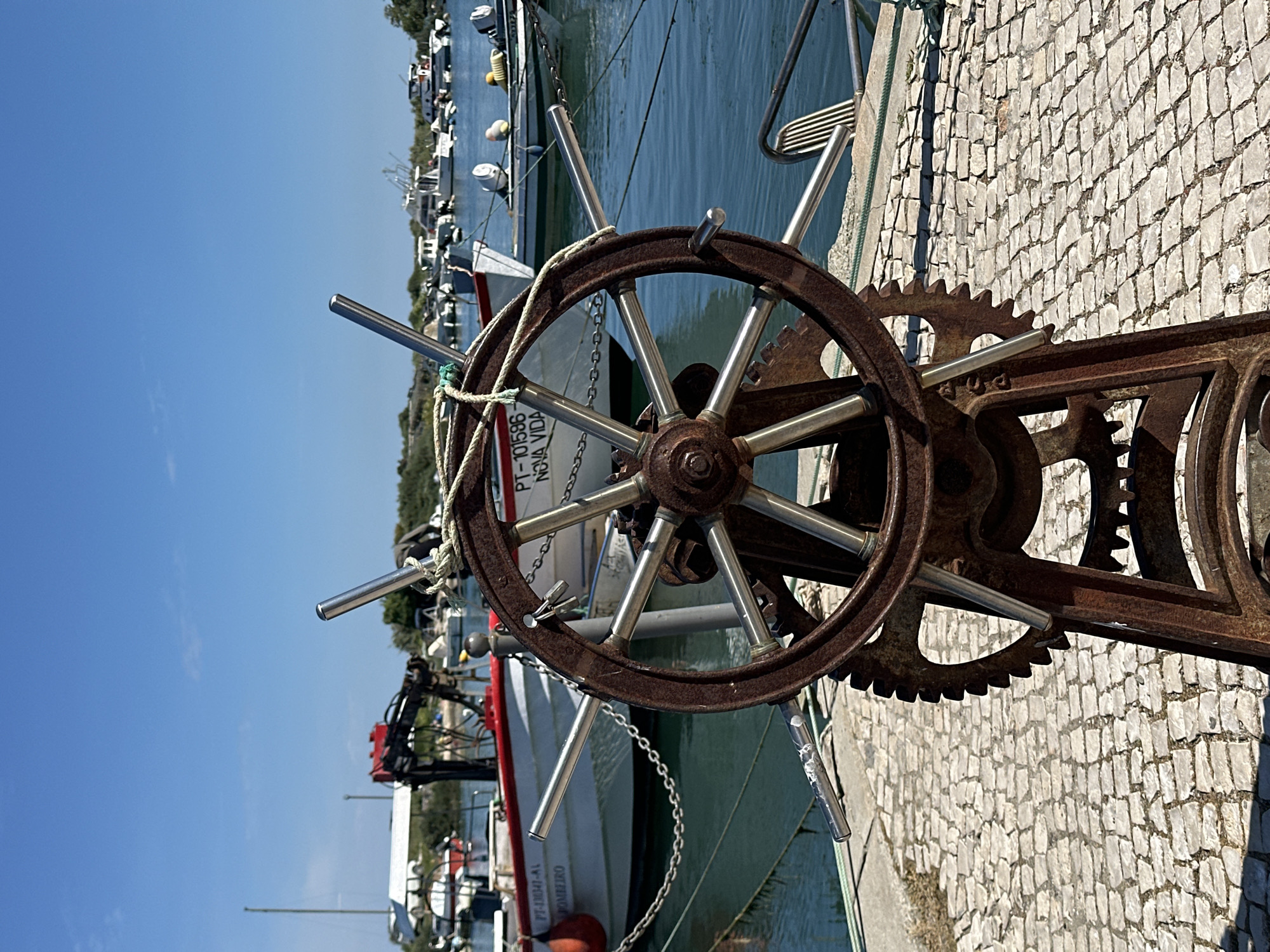 The image presents a close-up view of a rusty, metallic steering wheel apparatus situated on a paved surface. The steering wheel features a circular rim with metal rods extending from the center. A corroded gear mechanism is attached to the right side of the wheel.
In the background, a body of water is visible, populated with boats. One of the boats is partially visible, bearing the inscription "PT-101586 NOVA VIDA." The boat is painted white with a red stripe near the top of the hull.
The overall setting suggests a marina or waterfront location. Considering the text on the boat, it can be inferred that the location is in Olhão, Portugal.