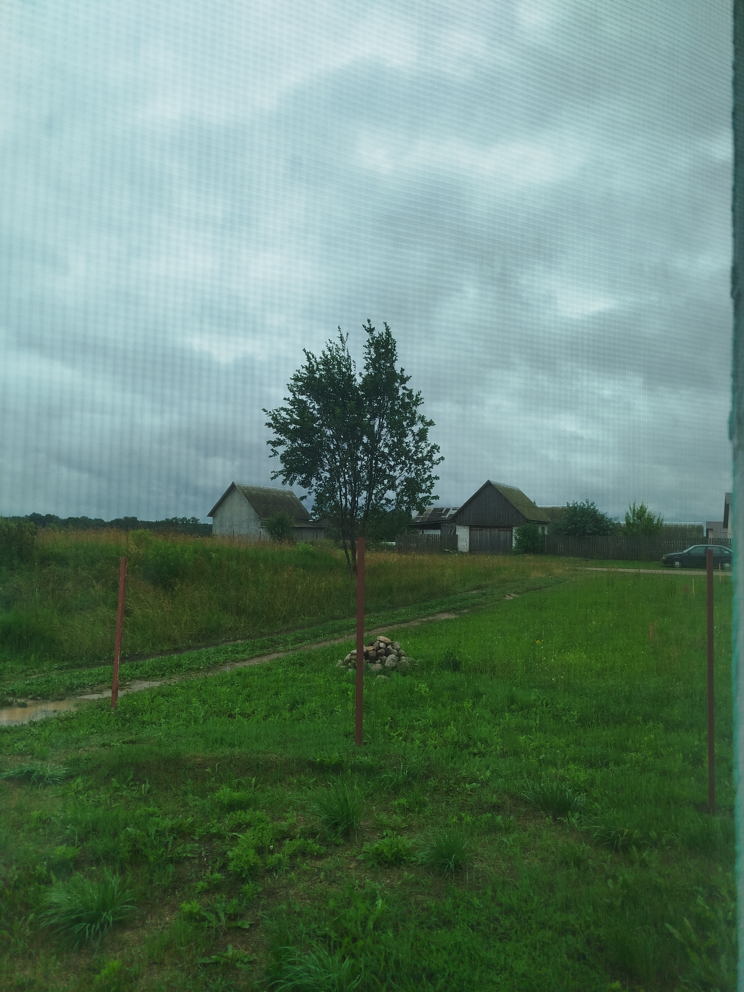 The image depicts a cloudy, overcast sky over a rural landscape. In the foreground, there is a grassy yard with two red posts marking a boundary. A dirt path runs through the grass. In the middle ground, there is a lone tree and two houses with dark roofs. The houses appear to be of simple construction. In the background, there are more trees and a fence. A dark-colored car is parked to the right of the scene. The overall impression is one of a quiet, somewhat somber rural setting, possibly after or during rain, as suggested by the overcast sky and a patch of damp earth on the path.