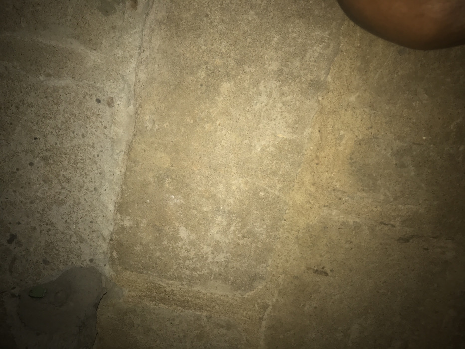 The image depicts a textured, beige-toned wall surface. Varying shades of tan and gray are present across the composition. The left section exhibits a rougher, more porous texture, while the center and right areas show a smoother, slightly weathered appearance. A curved, dark brown object occupies the upper right corner, potentially indicating an adjacent structure or feature. The lighting is uneven, casting shadows and highlighting surface irregularities.