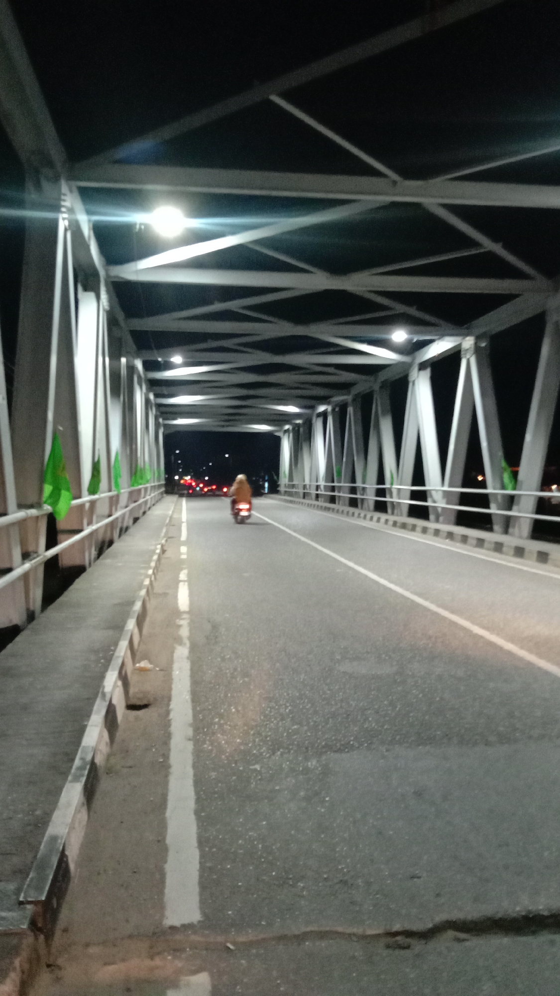 A nighttime scene shows a person riding a scooter across a bridge. The bridge's steel frame is visible overhead, with evenly spaced lights illuminating the roadway. Green flags are attached to the railing on the left side of the bridge. A white line marks the center of the road. At the far end of the bridge, blurred taillights of other vehicles can be seen. The location is Lhokseumawe, Indonesia.