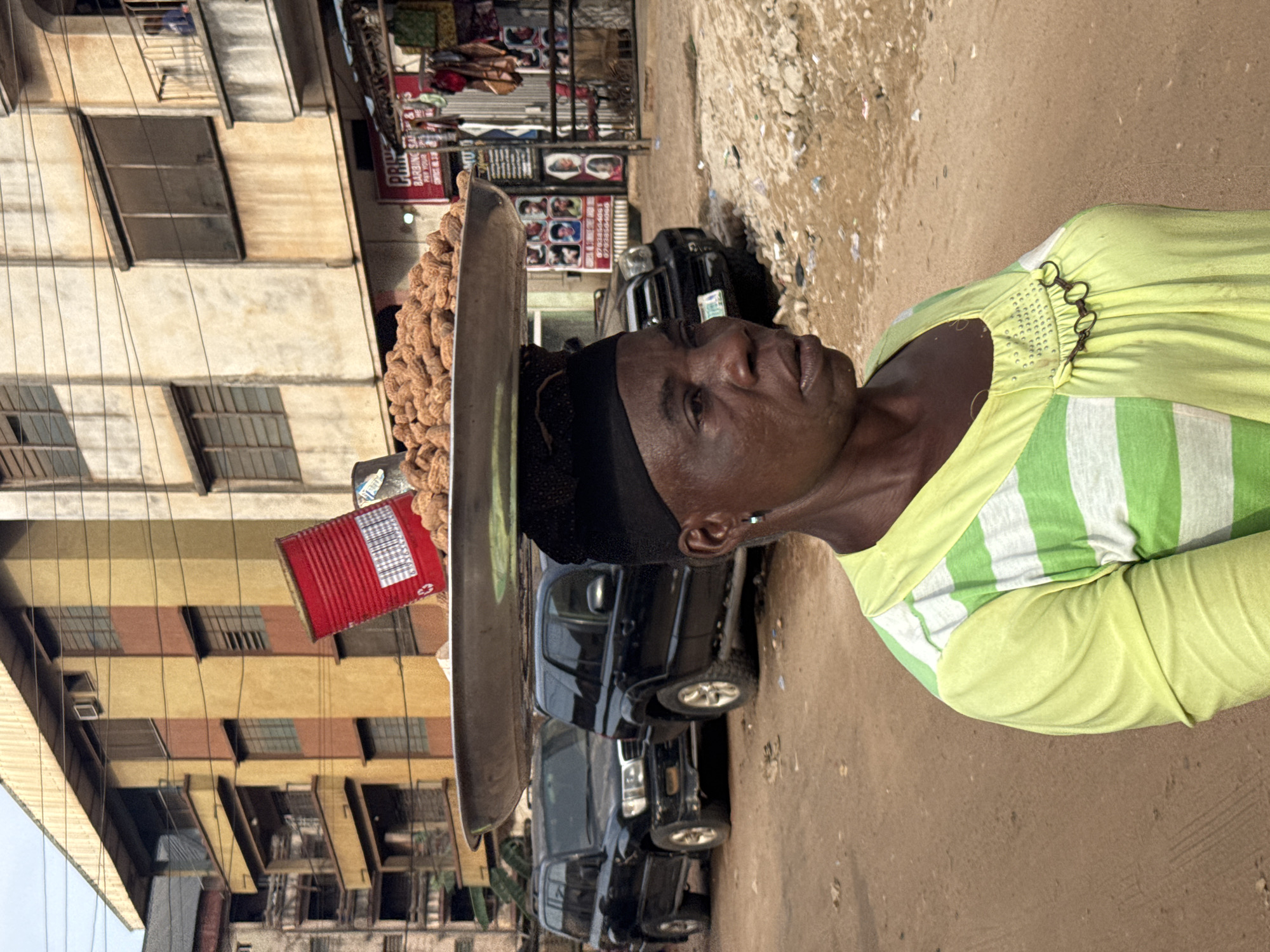An elderly woman in Onitsha, Nigeria, balances a large tray of peanuts on her head. The tray also has a red tin attached. She wears a black head covering and a lime green and white striped shirt. Several dark SUVs are parked behind her. The background includes multi-story buildings, some with visible store signs. The ground is dirt.