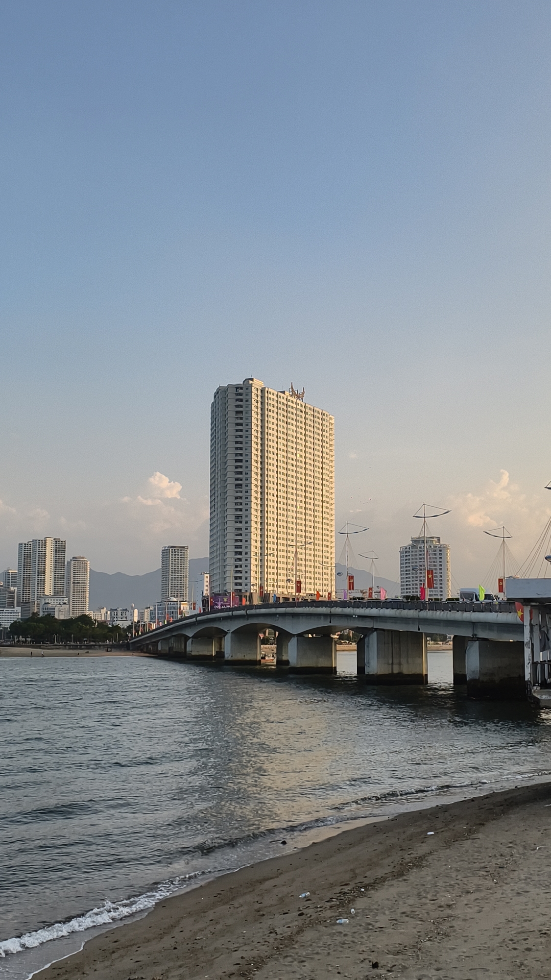 An outdoor, vertical, wide-angle shot captures a daytime view of a coastal city skyline. In the foreground, a sandy beach meets calm, dark water with gentle waves lapping at the shore. A concrete bridge spans across the water in the midground, its white underside and piers visible. Along the bridge, there are several tall buildings, including a prominent, light-colored skyscraper in the center, illuminated by the sun. Other buildings of varying heights line the cityscape beyond the bridge. In the background, mountains are visible in the distance. The sky is a clear, light blue with a few scattered white clouds. The overall scene suggests a modern urban environment situated by the sea.