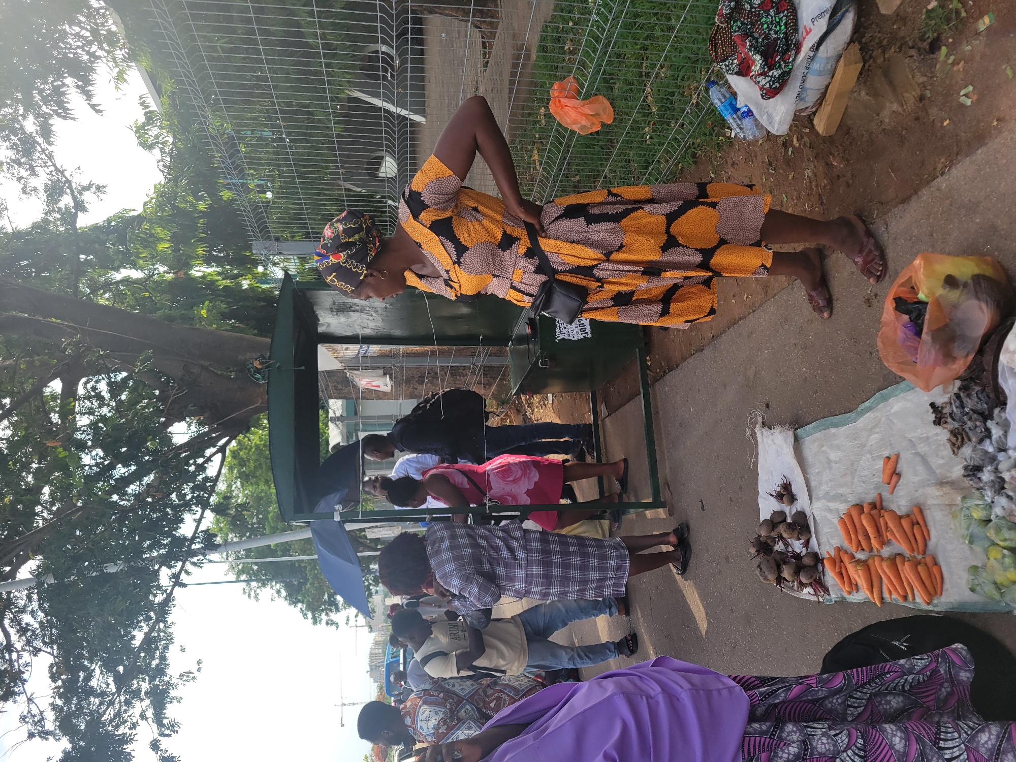 The image shows a street scene in Abuja, Nigeria. There is a line of people waiting at a small green kiosk. A woman wearing a yellow and black patterned dress and a patterned headscarf stands to the side. She has a black bag around her waist. Vendors are selling goods on blankets on the ground. These include beets, carrots and other produce. Other vendors also have goods in bags near the fence. People are standing and looking at what they want to buy. The sky is overcast.