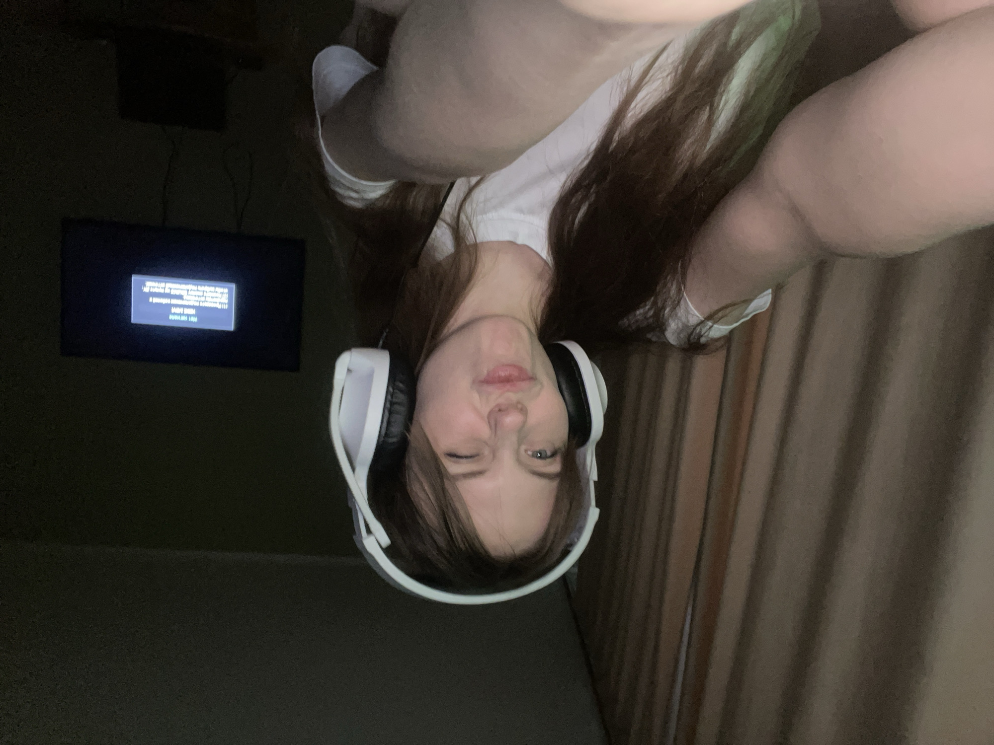 A young woman wearing a white t-shirt and white headphones is centered in the frame, turned upside down and winking. In the background, a television screen displays white text on a black background, positioned against a dark wall. A darker object is situated above the television. A curtain is on the right side of the scene, and the floor is visible to the left. The location is presumed to be Ufa, Russia.