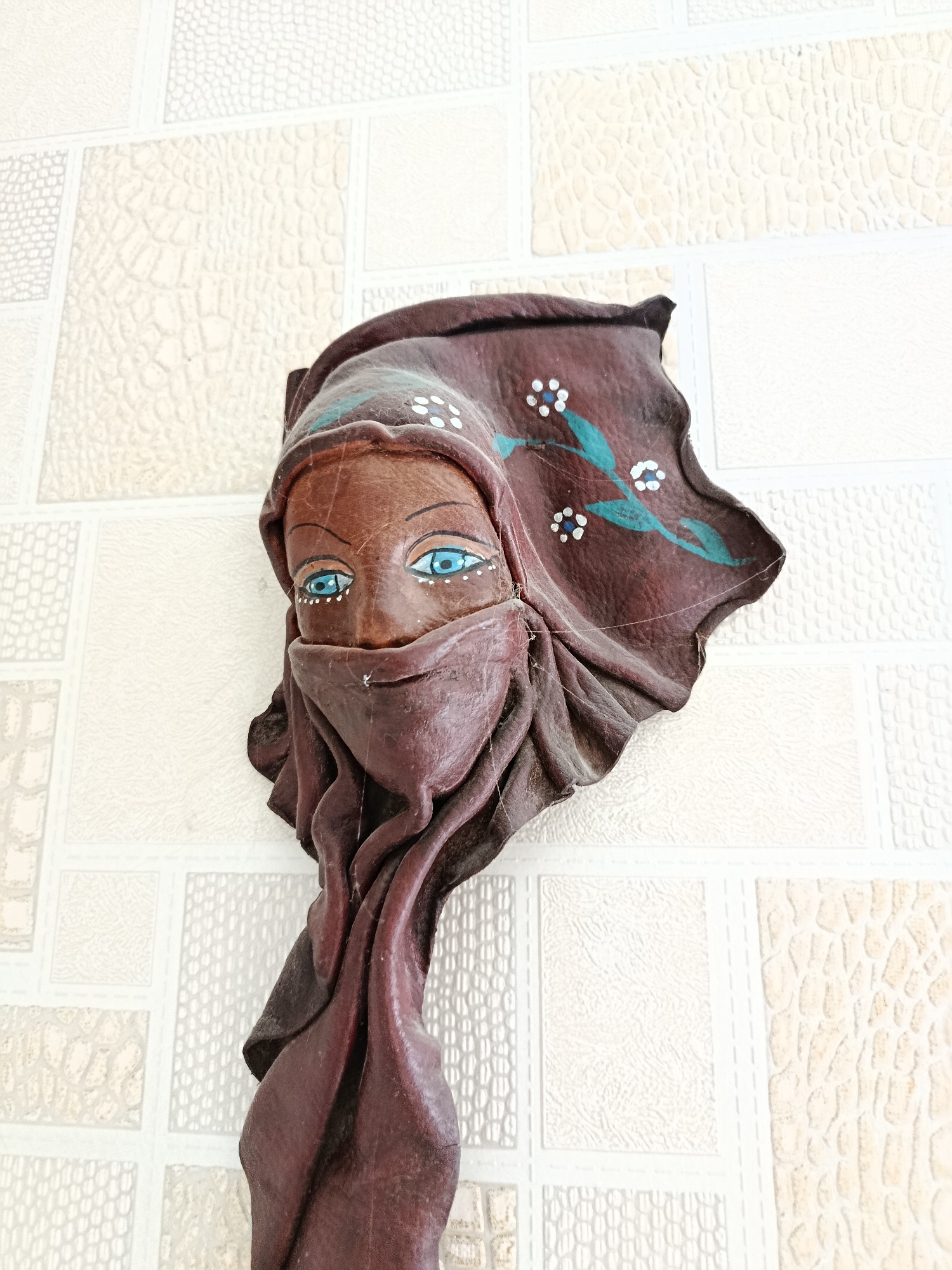 A wooden mask of a woman's face, wearing a headscarf and veil, is mounted on a textured, light-colored wall with a grid pattern. The mask is carved and painted with blue eyes and floral designs on the headscarf. The wall appears to be an interior surface, possibly in a home or establishment. The location is not specified within the image or its metadata, therefore, no information can be provided regarding its presence in Saratov, Russia.