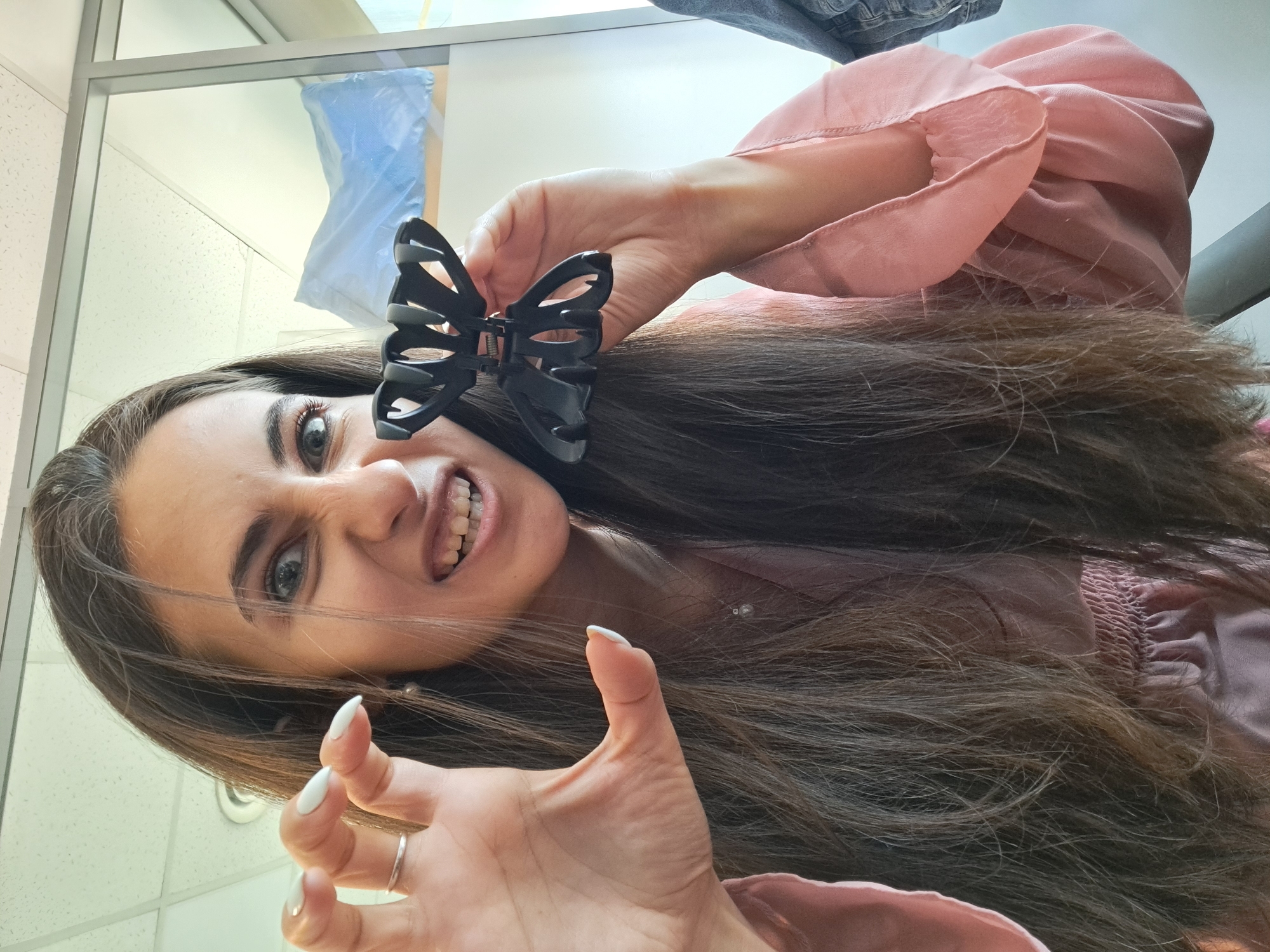 A young woman with long, dark brown hair is depicted in a playful, low-angle portrait, with the image rotated clockwise. She looks directly at the camera with wide, light-colored eyes and a mischievous expression, revealing her teeth in a grin. Her left hand is raised in a claw-like gesture, showcasing white, manicured stiletto nails and a silver ring on her ring finger. Her right hand holds a black, butterfly-shaped hair claw near her head. She is wearing a light pink top or dress made of a sheer, ruffled fabric, with smocked details visible beneath her hair. A delicate silver necklace with small beads adorns her neck. The setting appears to be indoors, likely an office or commercial building, indicated by white ceiling tiles and a window or glass partition in the background. A light blue object, possibly fabric or a bag, is reflected in the glass, along with what might be denim clothing hanging. The overall ambiance is energetic and theatrical. No visible text is present. The location context is Sofia, Bulgaria.