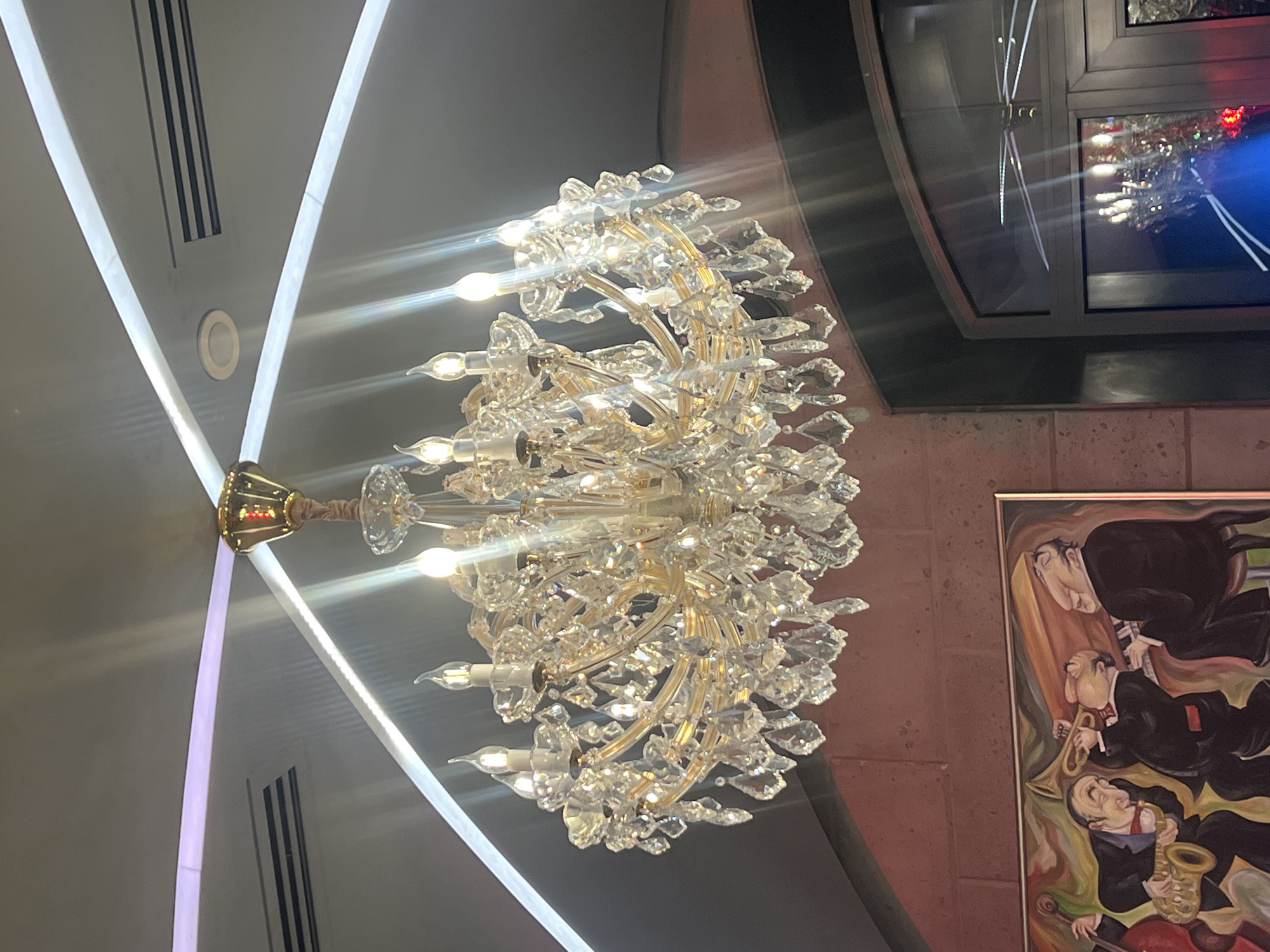 A crystal chandelier hangs from the ceiling in Yerevan, Armenia. The chandelier is gold with multiple layers of crystals. There is a painting of three jazz musicians in the lower right corner of the image. There is a window in the upper right corner of the image reflecting light.