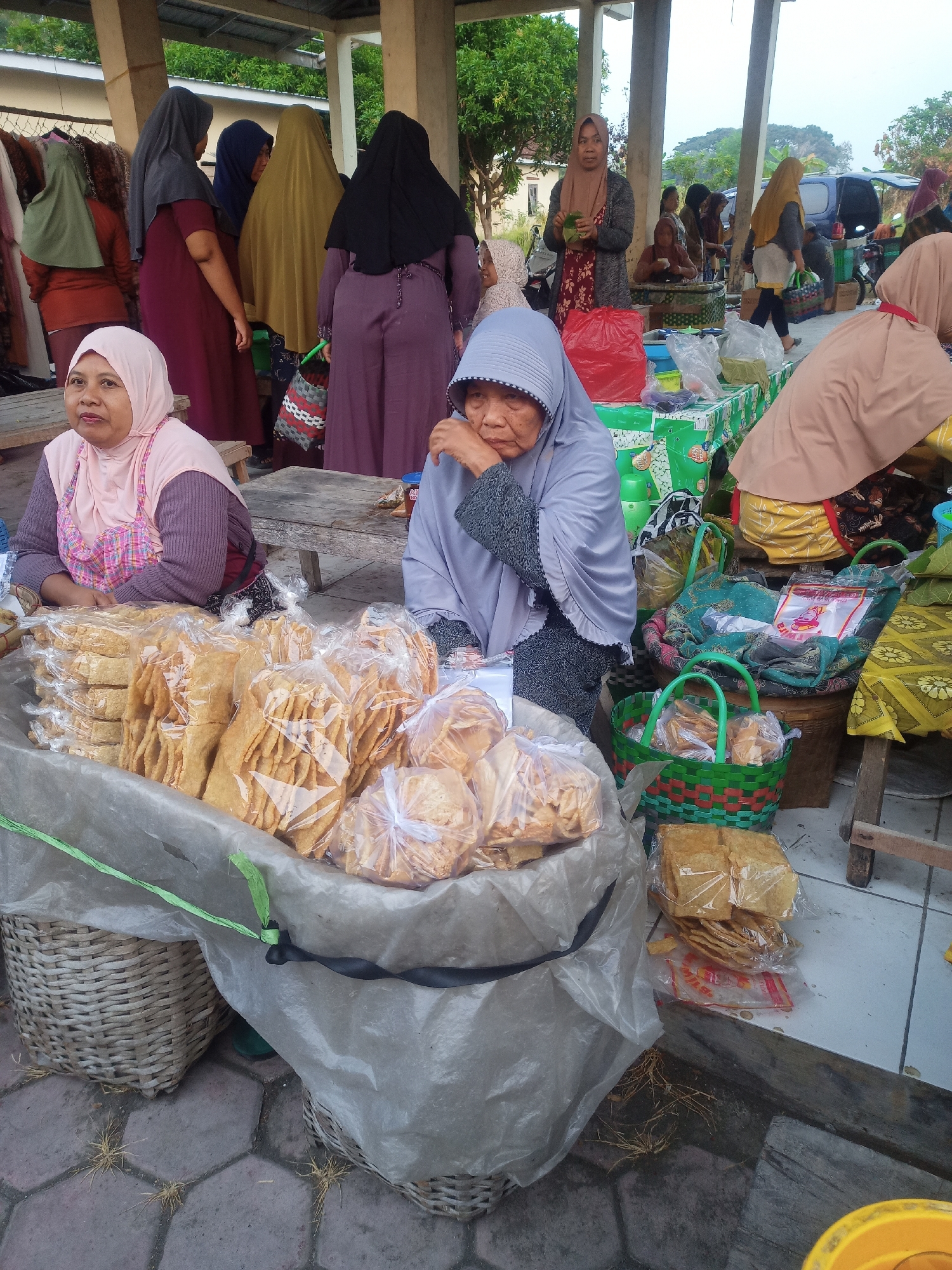 In the city of Nganti, Indonesia, a group of women are gathered around market stalls, likely in an outdoor setting. The foreground is dominated by a large display of packaged crackers and snacks, presented in plastic bags and a large woven basket lined with clear plastic. Two women are seated behind this display. The woman on the left wears a pink hijab and a purple patterned shirt, looking towards the viewer. The woman on the right wears a light blue hijab and a dark patterned top, with her hand touching her face.
Behind them, other women are visible, some in colorful hijabs and traditional attire, interacting with each other and tending to their goods. One woman in a dark purple abaya stands with her back to the camera. Another, in a mustard yellow hijab, is walking away from the viewer. The market appears to be under a covered structure, with concrete pillars and a corrugated roof visible. Tables and baskets are laden with various items, and in the background, trees and some parked vehicles can be seen, suggesting a bustling marketplace environment. The ground is paved with hexagonal tiles.