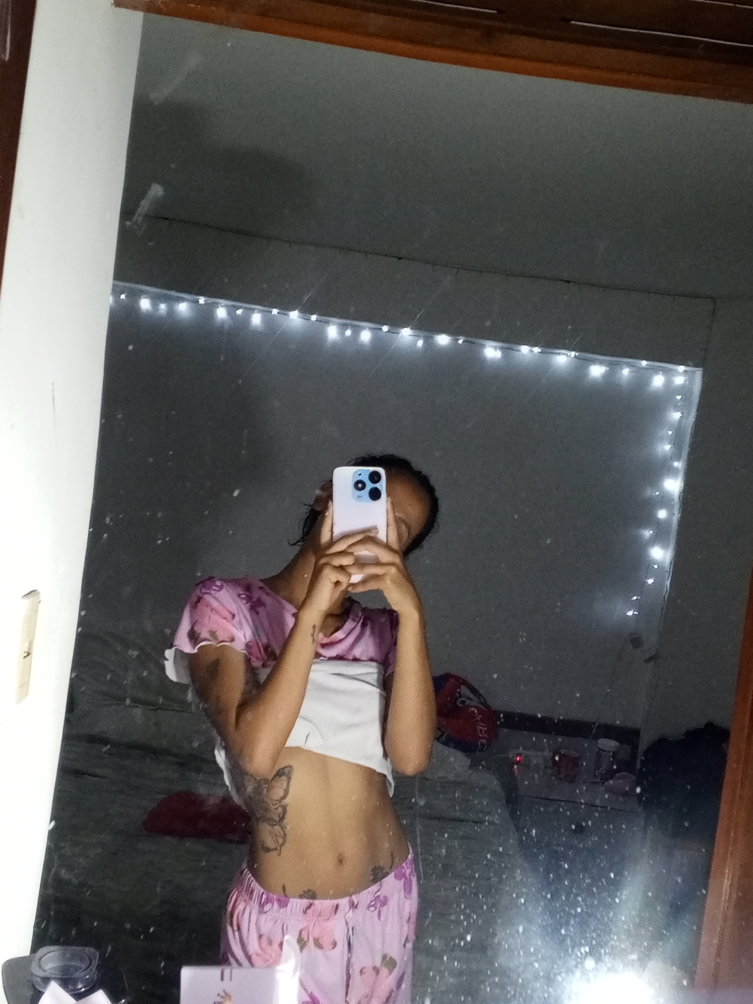 A person is captured in a mirror selfie, holding a light-colored smartphone with a triple-lens camera system that obscures their face. They are wearing a short-sleeved, cropped top with pink and white patterns, revealing their midriff, and loose-fitting pink patterned shorts. Visible on their left rib cage is a tattoo depicting a butterfly, and another smaller tattoo is on their lower abdomen.
The mirror itself is speckled with numerous small particles, appearing like dust or light reflections. It is framed by a wooden border on the top and right sides. The background reflected in the mirror shows a dimly lit room, likely a bedroom, with white string LED fairy lights draped along the top and right edges of the reflected wall, providing illumination.
Reflected in the background are glimpses of a bed with white bedding, where a red bag or object and other dark items are visible. To the bottom left of the reflection, a container with a dark lid sits on a surface. A white light switch is present on the wall to the left of the mirror's edge. The overall impression is an intimate, casual moment, likely occurring indoors during the evening or night, in a location with ambient lighting provided by fairy lights. No legible text is visible in the image.