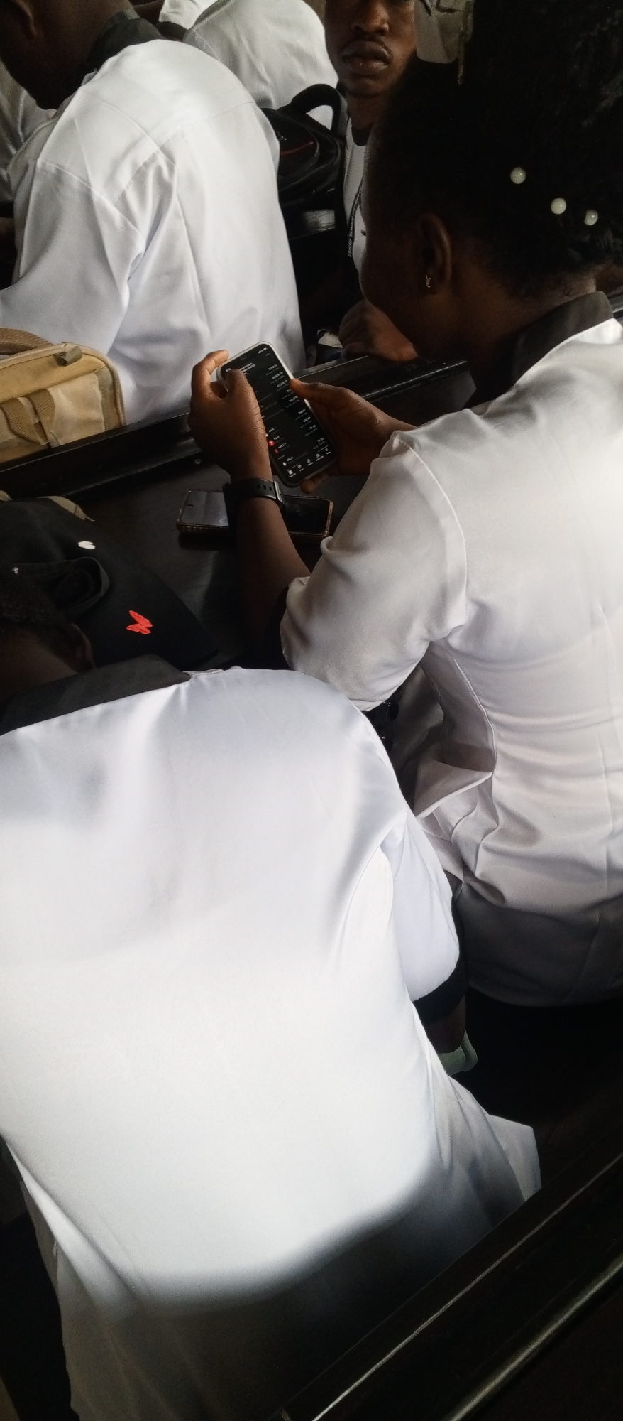 A medium shot captures a group of people in what appears to be a classroom or lecture hall setting in Kuson, Nigeria. The individuals are dressed in white lab coats, suggesting a medical or scientific context. The foreground is dominated by the back of a person wearing a white lab coat, their head bowed. To the right, another person in a white lab coat is looking down at a smartphone held in their hands. The phone screen displays a financial chart or trading platform. Other individuals in white lab coats are partially visible in the background, some with their heads turned, others looking forward. The environment is dimly lit, with the dark surfaces of tables and chairs contrasting with the white attire of the people. A beige bag is visible on the left side of the frame. The overall atmosphere appears serious and focused, with the individuals engaged in their respective activities.
