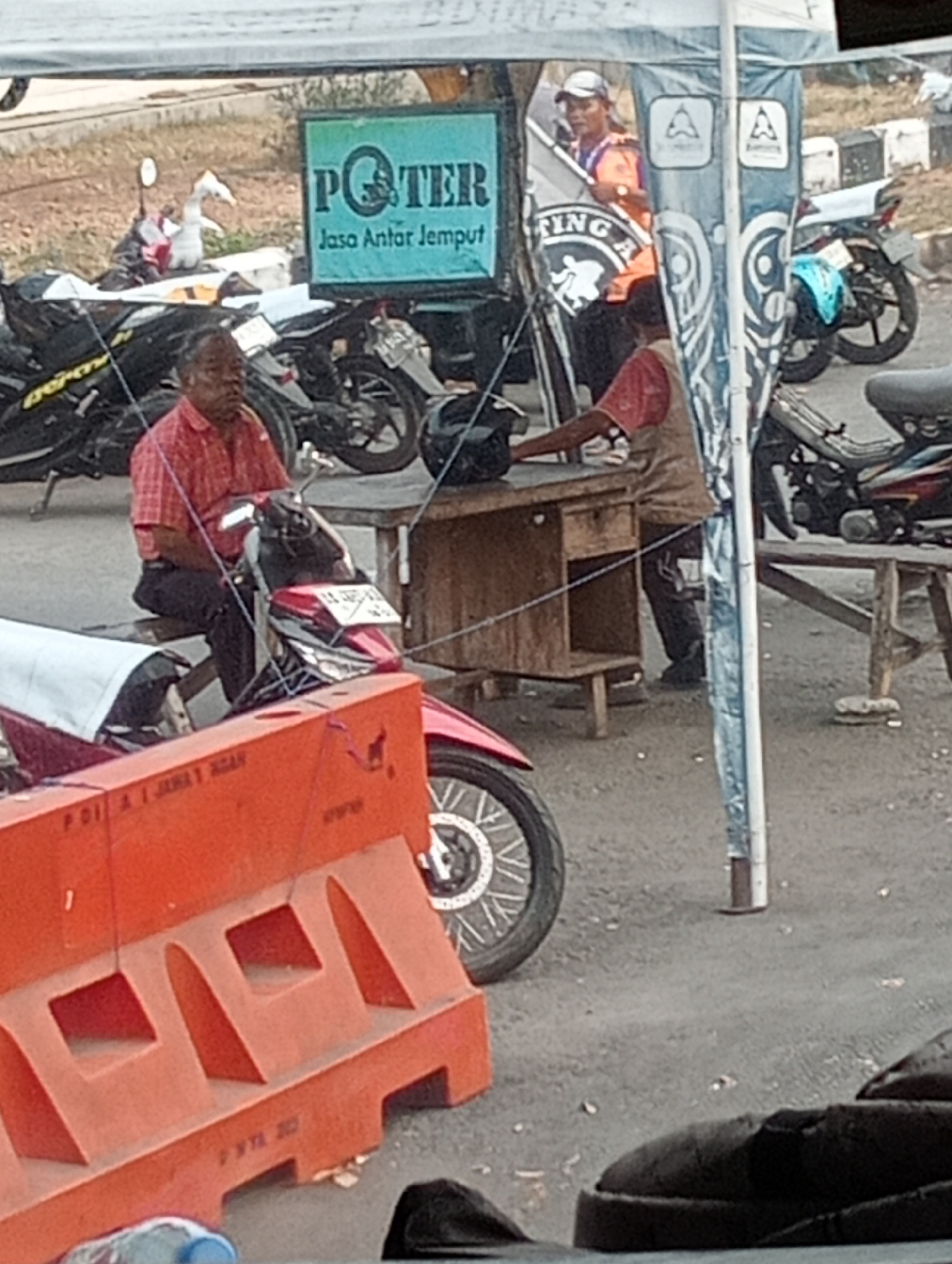 The image depicts an outdoor scene, likely in Magelang, Indonesia, with a focus on a temporary setup involving motorcycles and a canopy. In the foreground, a man in a red checkered shirt and dark pants is seated on a red and black motorcycle. He appears to be looking to his left, with his hands resting on the handlebars. To the left of the man, an orange construction barrier is visible, with some writing on its surface.
Behind the man, a wooden table is set up, with a black helmet placed on it. A person wearing an orange vest and a cap is standing behind the table, looking towards the left of the frame. Several motorcycles are parked in the background, some with visible license plates. One motorcycle has a blue fender. A green sign with the word "POTER" and text below it, possibly indicating a service, is also visible behind the parked motorcycles. The ground is paved with asphalt. The overall impression is of a roadside or market area where motorcycle-related services might be offered.