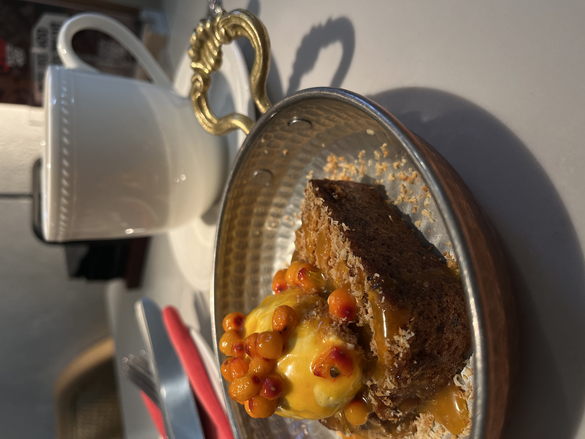 A close-up shot shows a slice of cake with sea buckthorn berries and sauce in a metal dish with a gold handle. To the left, partially visible, is a white teacup on a saucer. Silver cutlery with a red handle is partially visible on the left. The scene appears to be indoors, with light coming from the upper right, casting shadows. The exact location in the city of Krasnoyarsk, Russia, cannot be determined from the image.