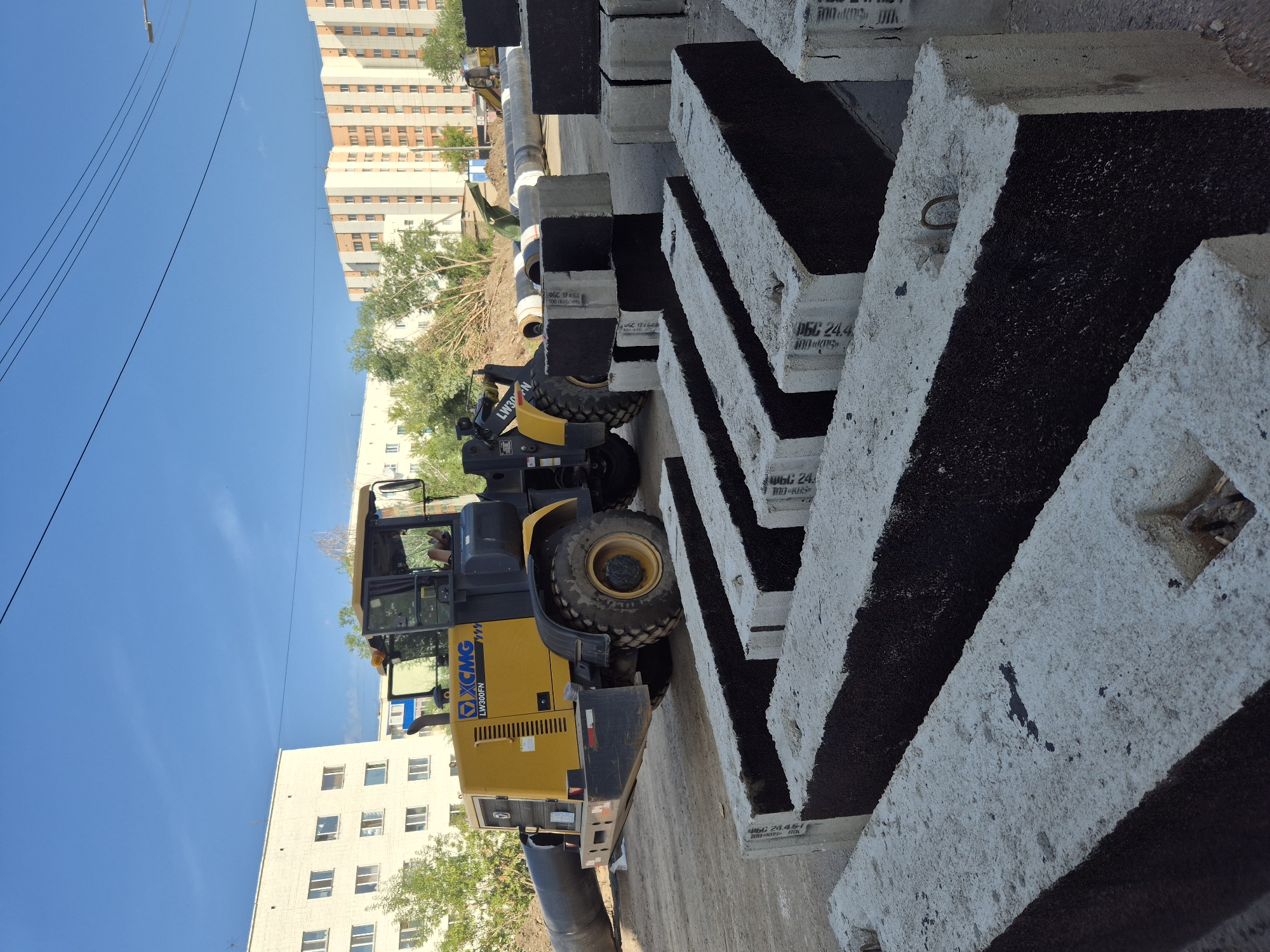 A construction site in Kokshetau, Kazakhstan, is depicted under a clear blue sky. In the foreground, several large, rectangular concrete blocks with dark inserts are neatly arranged. Behind them, a yellow XCMG LW300FN wheel loader is parked. The loader has a large black pipe extending from its front. In the background, two multi-story apartment buildings are visible, along with trees and more construction materials including large pipes. The scene suggests ongoing infrastructure work.