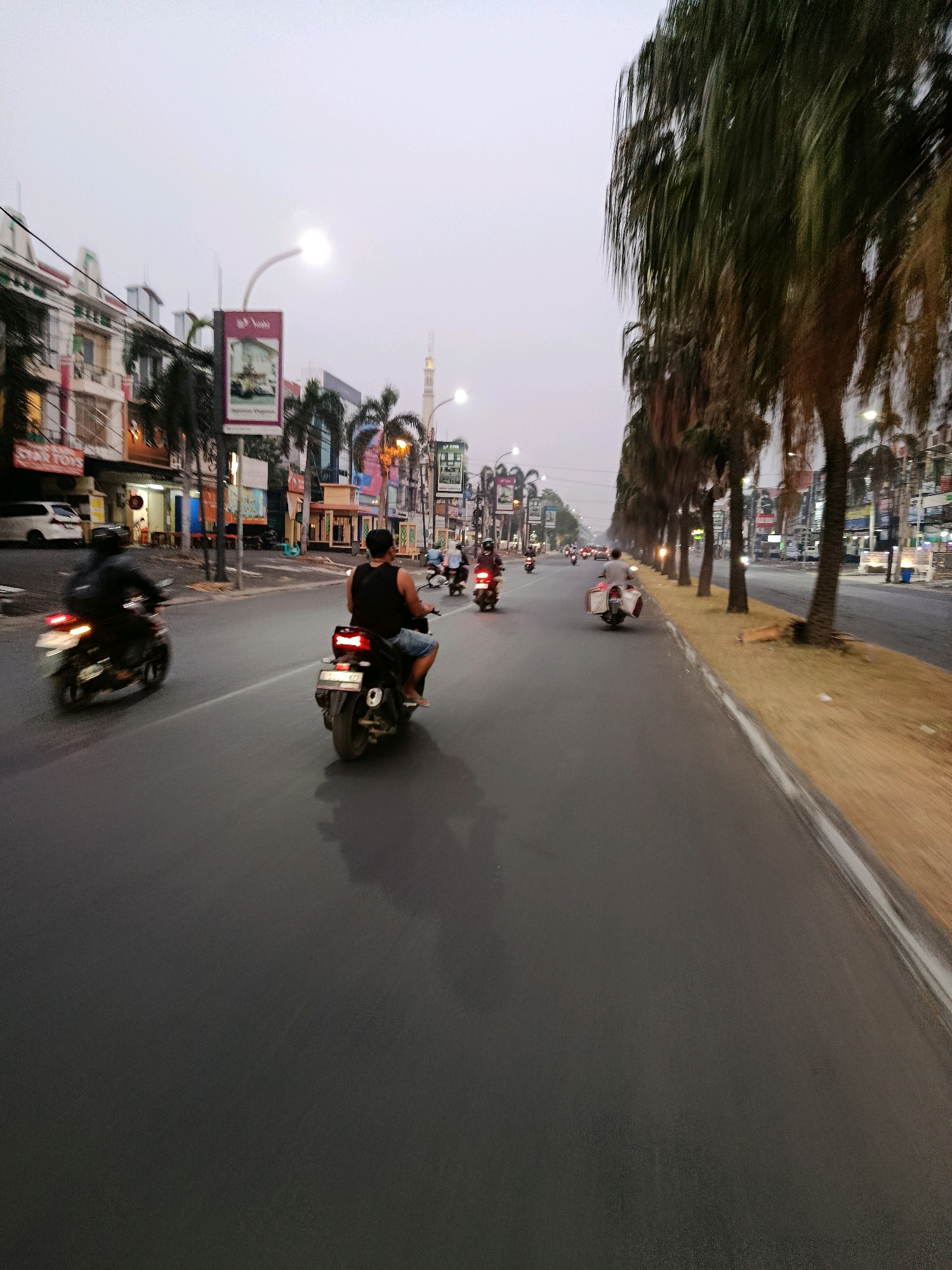 The image depicts a street scene in Bekasi, Indonesia, during twilight. Several motorcycles are visible on the asphalt road. In the foreground, a person wearing a black sleeveless shirt and blue shorts rides a motorcycle with a prominent rear brake light. To the left, another motorcycle with a rider is in motion, its taillight blurred by movement. Further down the road, other motorcyclists are also visible, some with passengers.
The street is lined with palm trees on the right side, with their fronds creating a dappled effect. On the left, buildings with shopfronts and signage are present, including a white van parked near one of the establishments. Streetlights illuminate the scene. In the background, a minaret of a mosque can be seen. The sky is overcast and transitioning from day to night. The overall impression is that of a busy urban street with ongoing traffic.
