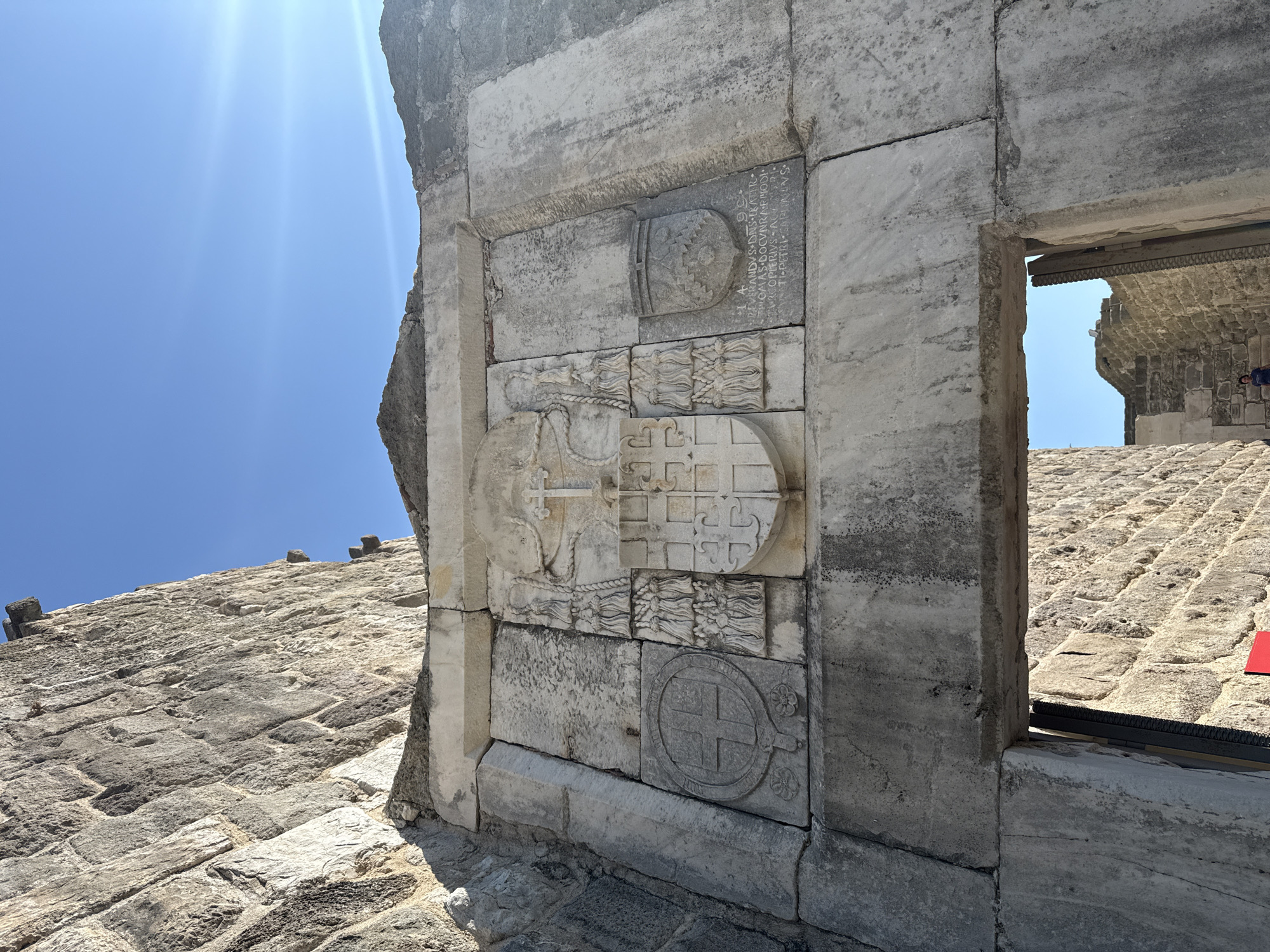 The image captures a close-up view of a stone archway in Bodrum, Turkey, likely part of the Bodrum Castle (Castle of St. Peter). The archway is framed by weathered, light-colored stone blocks. Above the arch, a large, intricately carved stone relief is prominent. This relief features a central heraldic crest with a cross at its apex, flanked by decorative elements. To the left of the crest is a circular carving with a cross inside and floral motifs at the bottom. To the right, a shield-shaped carving contains a more intricate pattern. Above the shield, some Latin inscription is visible. The stone surfaces show signs of age and wear. The sky above is a clear, bright blue with the sun casting a lens flare. Below the arch, through the opening, a stone-walled passage is visible, leading to a courtyard or interior space with more stone structures and a person walking in the distance. The overall impression is one of historical significance and architectural grandeur.