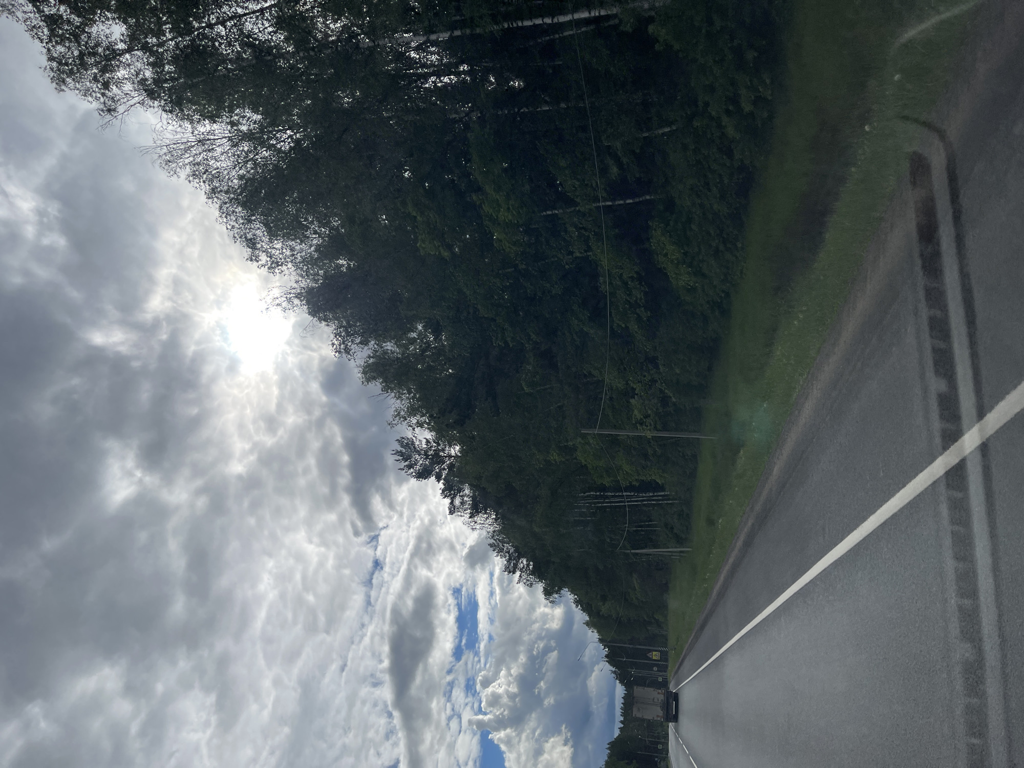 The image depicts a road running alongside a forest, presumably in the vicinity of Semyonovskoye, Russia. The road is asphalt, with a white painted dividing line visible. The forest consists primarily of trees with dense foliage. The sky is partly cloudy with a bright sun partially obscured by the clouds. The viewpoint is from within a vehicle, likely driving on the road.